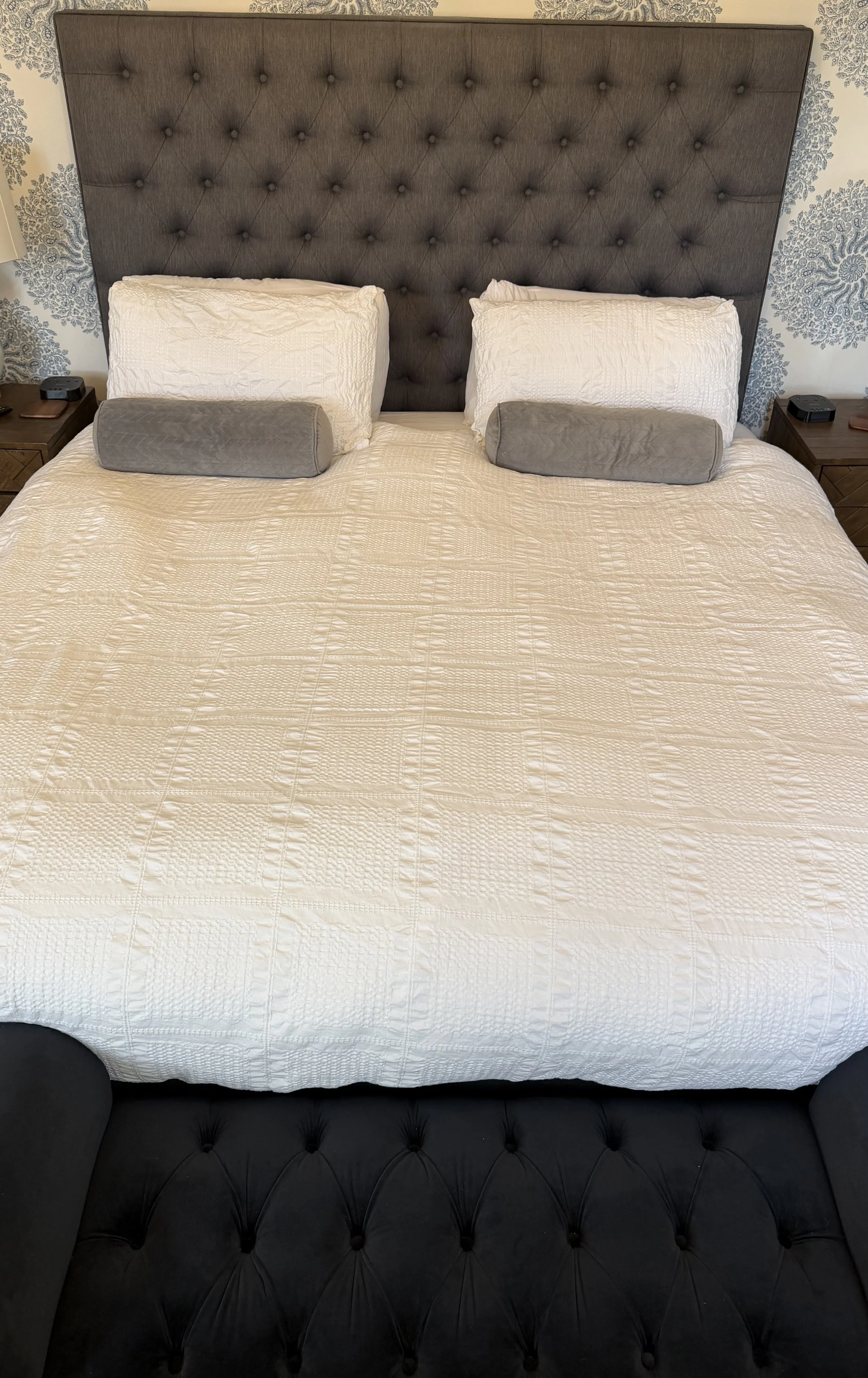 A neatly made bed with a beige quilt, two large white pillows, and two smaller gray cylindrical pillows, against a tufted dark headboard in a bedroom with patterned wallpaper.