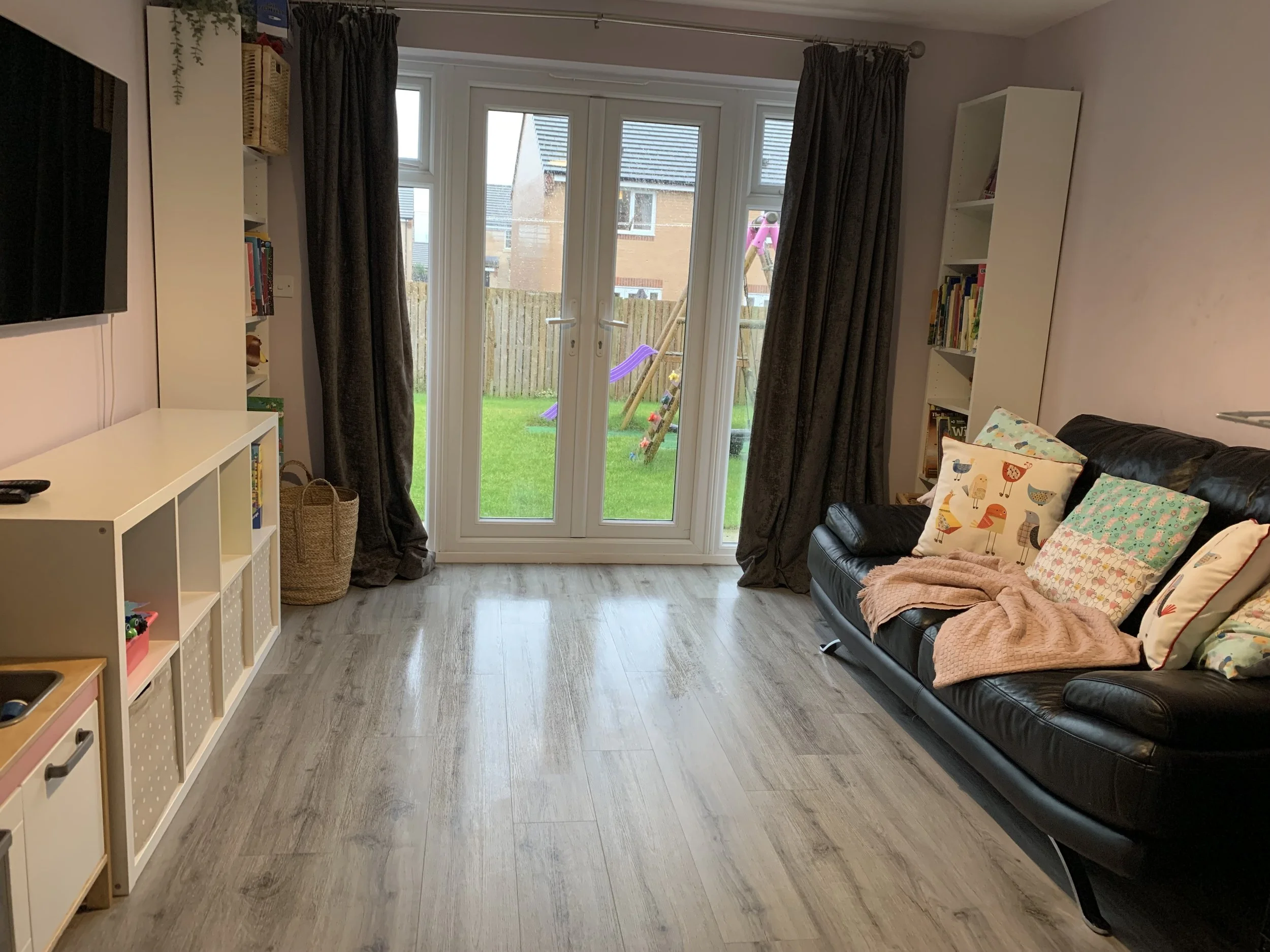 A living room with a glass door leading to a backyard, featuring a black leather sofa with colorful pillows, white bookshelves, and dark curtains. Outside is a grassy yard with a pink and purple swing set.