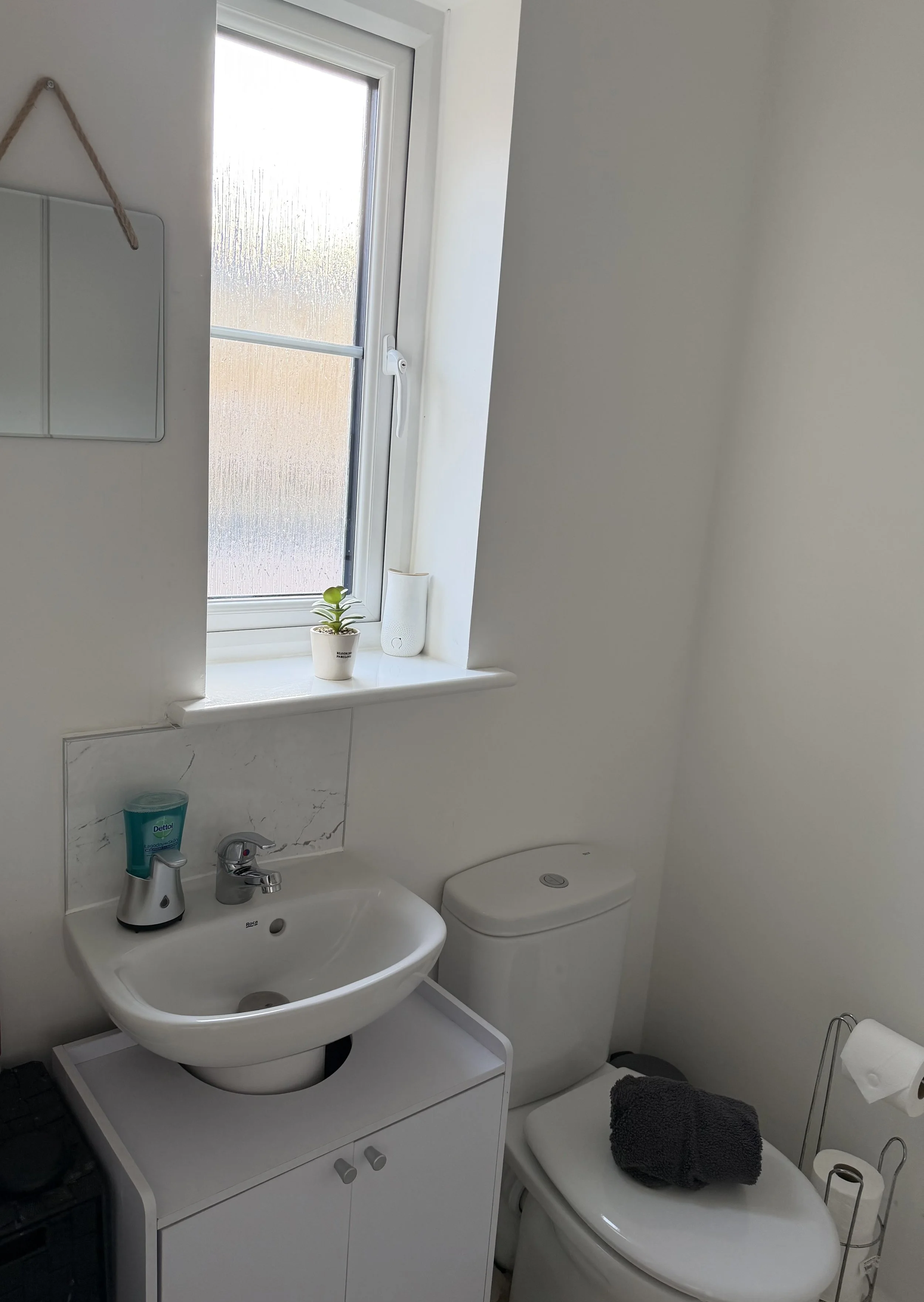 A small bathroom with a white sink, a toilet with a dark towel on the lid, a white cabinet under the sink, a window with frosted glass, a small potted plant on the window sill, a mirror on the wall, and a roll of toilet paper on a stand.