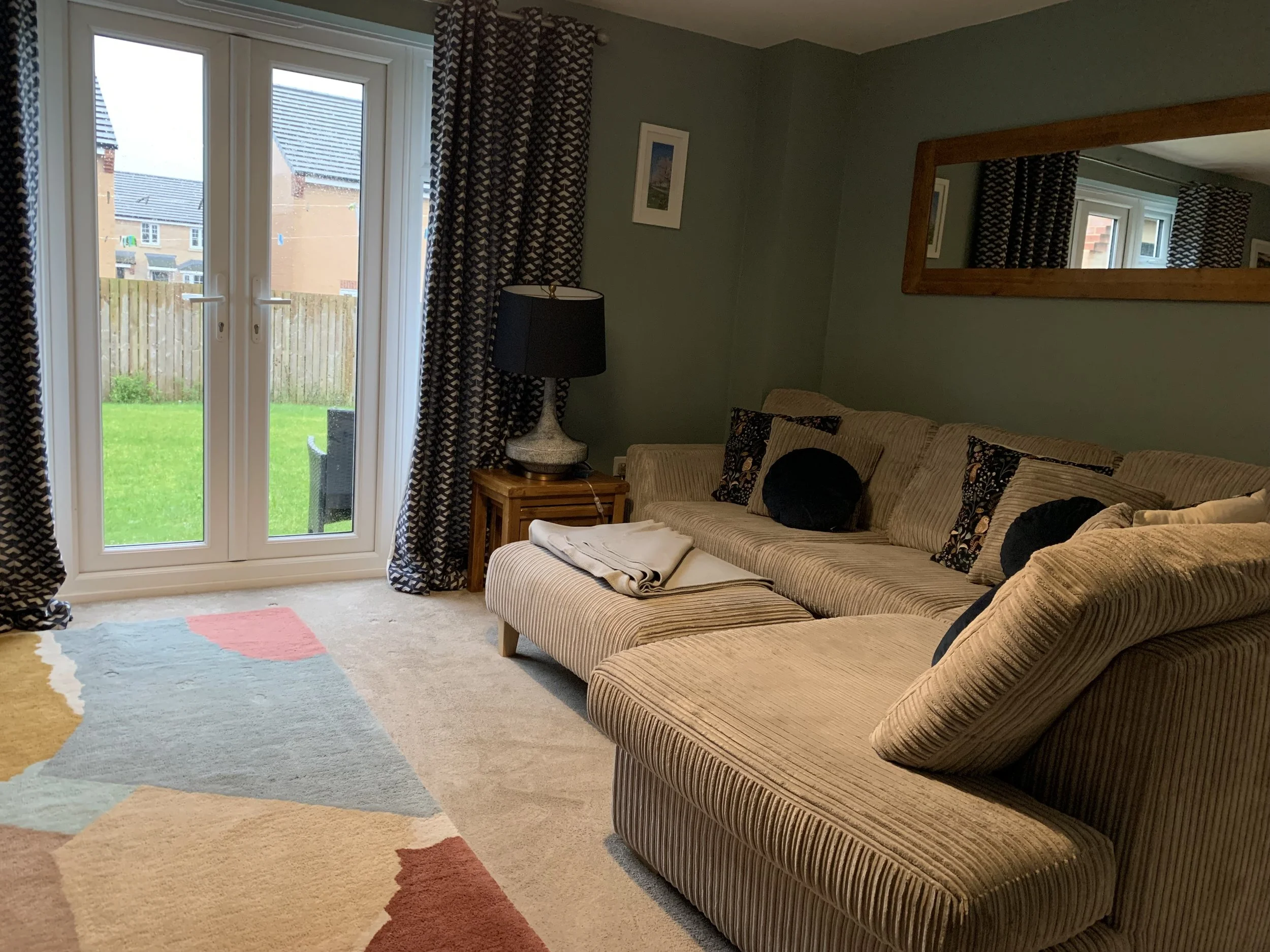 Living room with beige striped sectional sofa, decorative pillows, wooden side table with lamp, and large mirror on green wall. Glass double doors lead to a backyard with green grass and wooden fence.