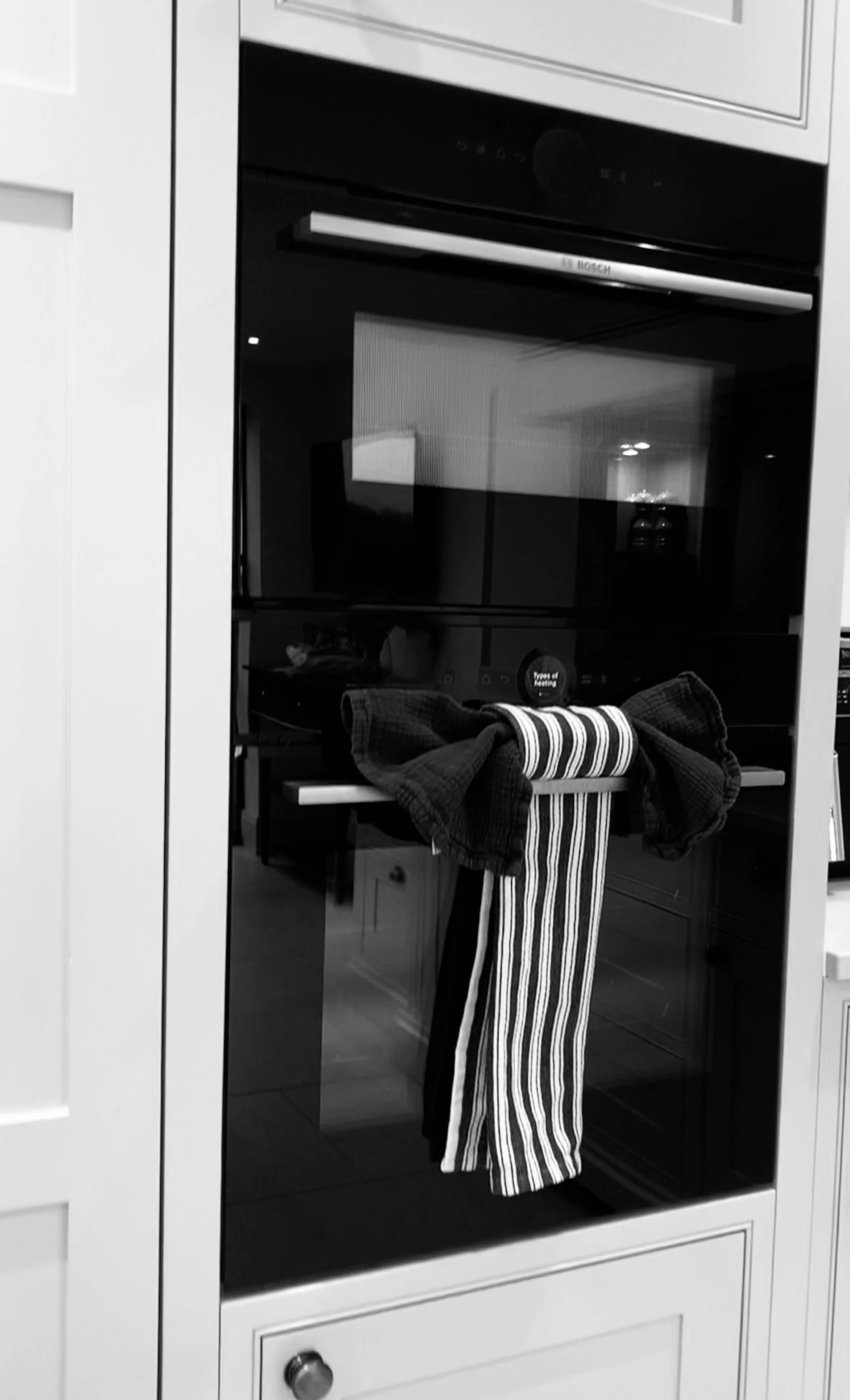 Black and white photo of a built-in oven inside kitchen cabinetry, with a cloth and striped dish towel hanging on the oven handle.
