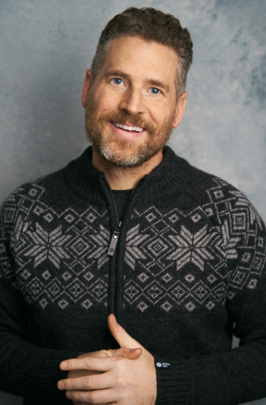 A smiling man with light skin, short curly dark hair, a beard, and blue eyes, wearing a black sweater with a white snowflake pattern.