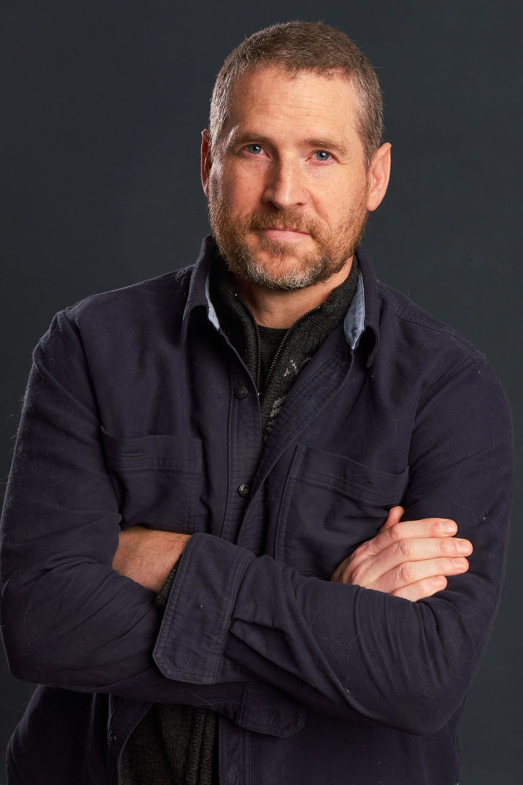 A man with short hair, a beard, and blue eyes, wearing a dark jacket, poses with arms crossed against a dark background.