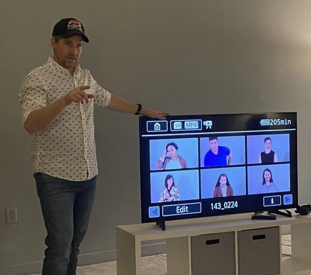 A man standing next to a flat-screen TV displaying a video call with nine participants.