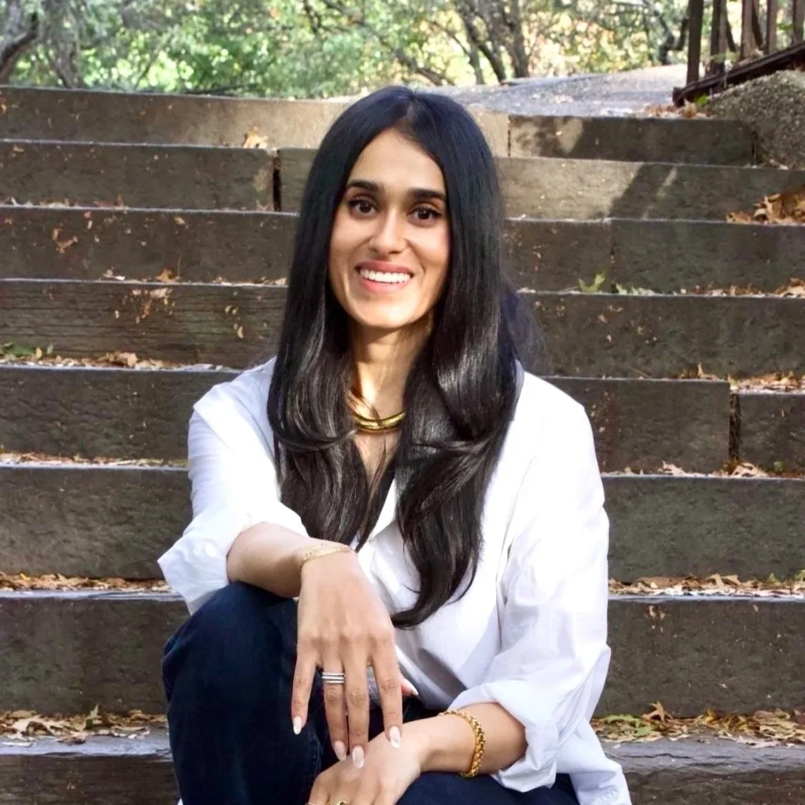 A woman with long black hair, wearing a white shirt, jewelry, and dark pants, sitting on the outdoor steps of a wooded area, smiling at the camera.