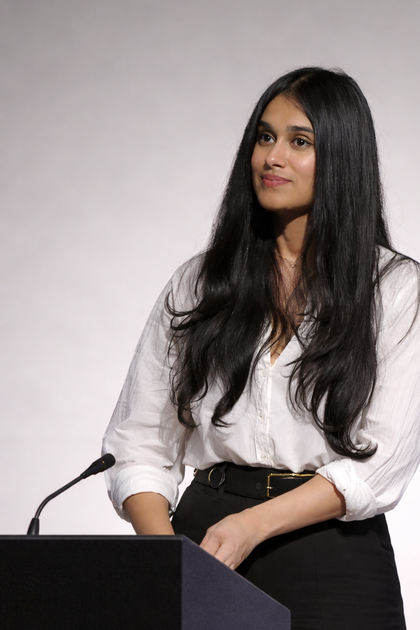 A young woman with long black hair, wearing a white shirt and black pants, stands at a black podium with a microphone, looking slightly to her right with a slight smile, against a plain gray background.