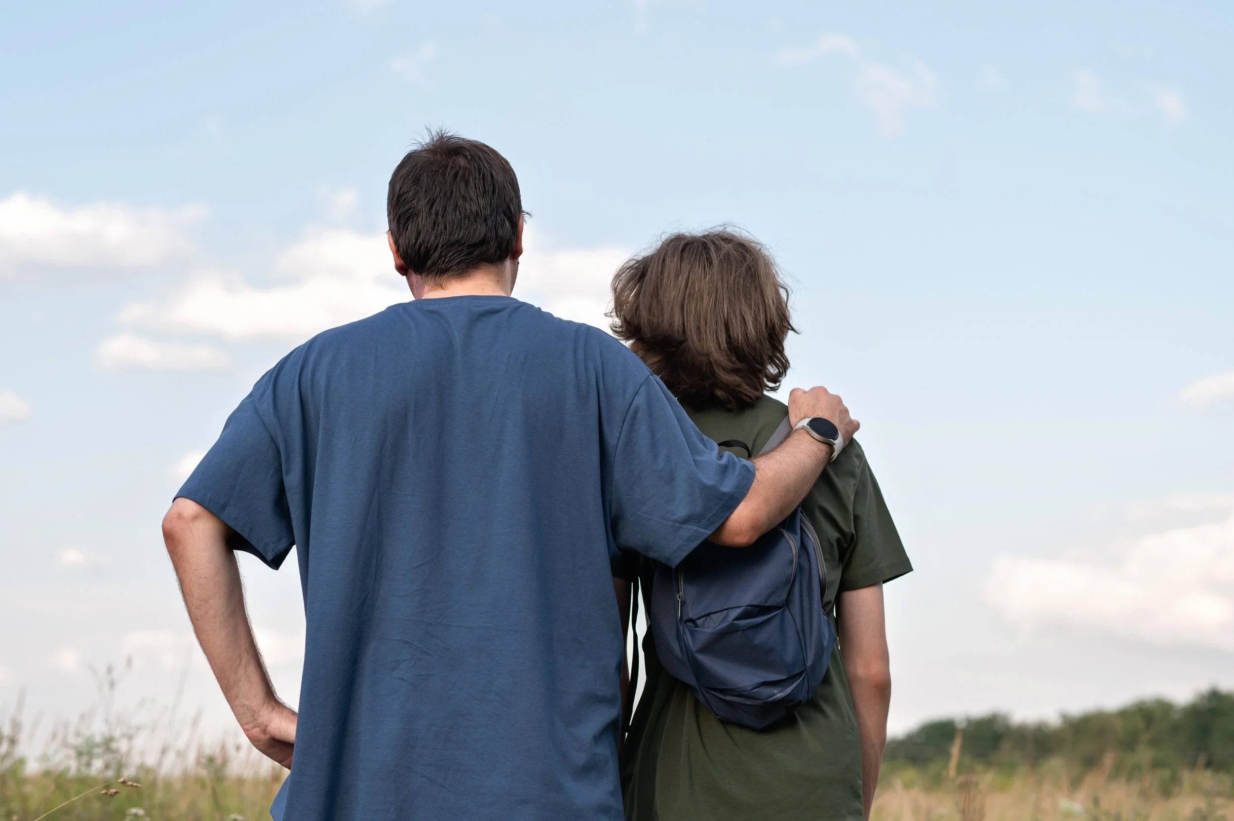 A man with a smartwatch on his wrist has his arm around the shoulder of a woman with a backpack as they look out over a grassy field with a few clouds in the sky.