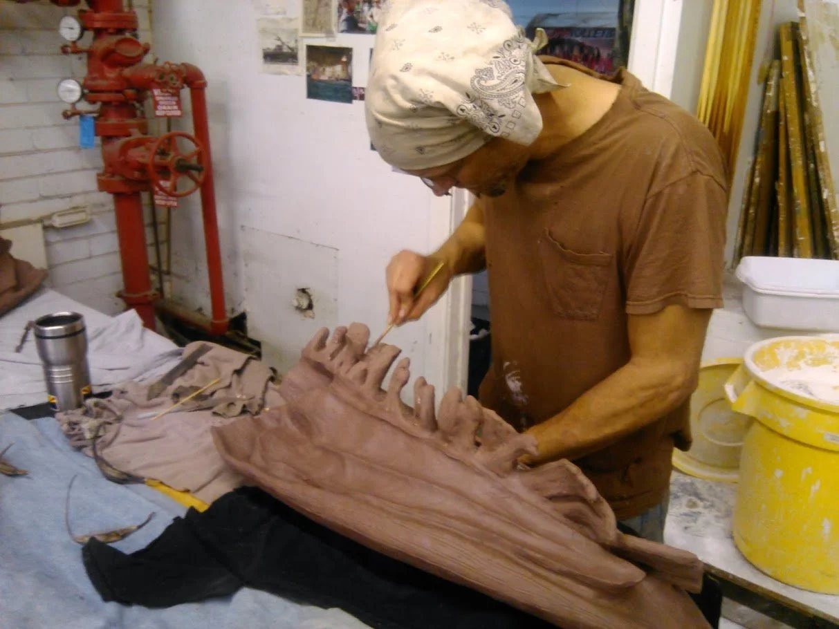 A person working on a large fish sculpture, possibly a whale, in an art studio. The person is wearing a bandana and a brown shirt, focusing on detailing the sculpture's teeth. The workspace has tools, paint buckets, and photographs on the wall.