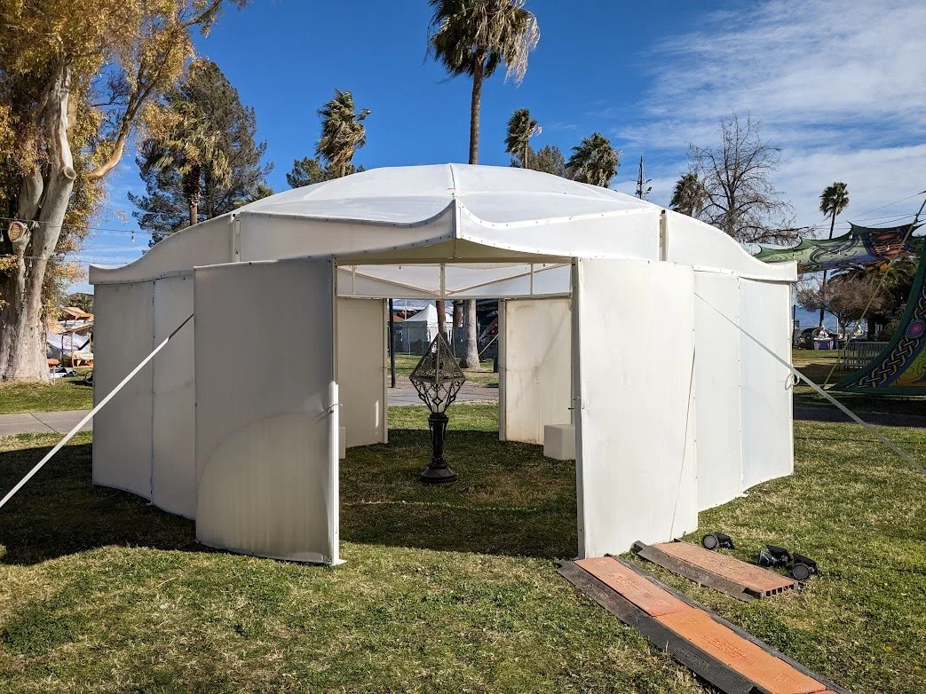 A white, semi-circular event tent on grass with open flaps in a park, surrounding trees, and a cloudy blue sky.