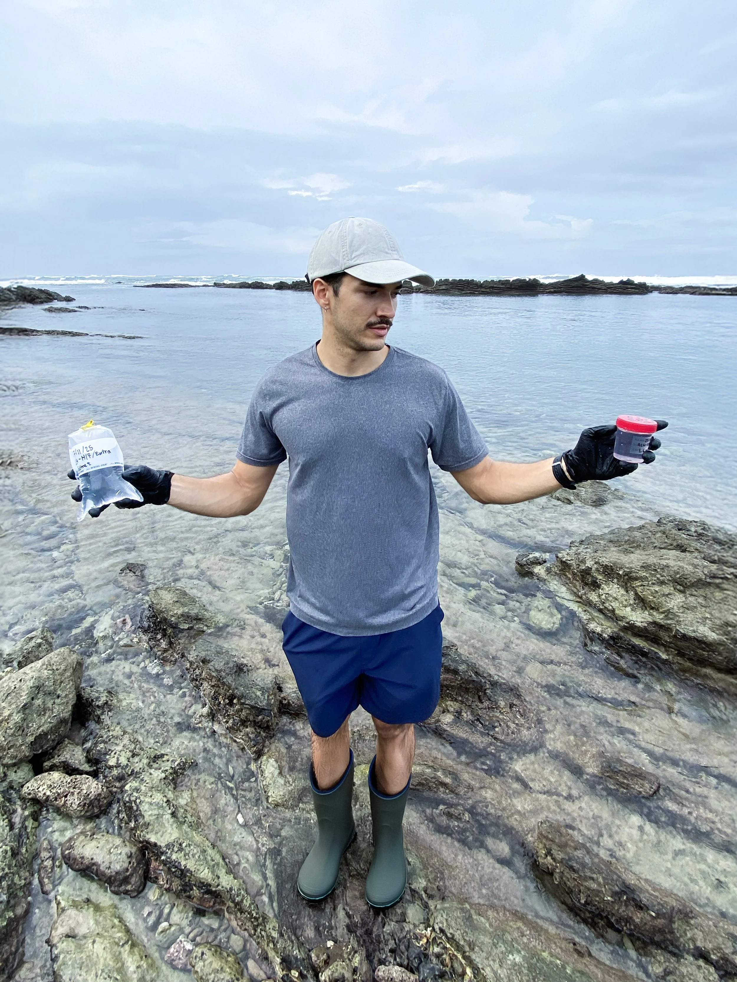 A man standing knee-deep in rocky water at the beach, holding a small container in one hand and a plastic bottle in the other, wearing a gray t-shirt, dark shorts, a light gray cap, and green rain boots, with ocean and cloudy sky in the background.