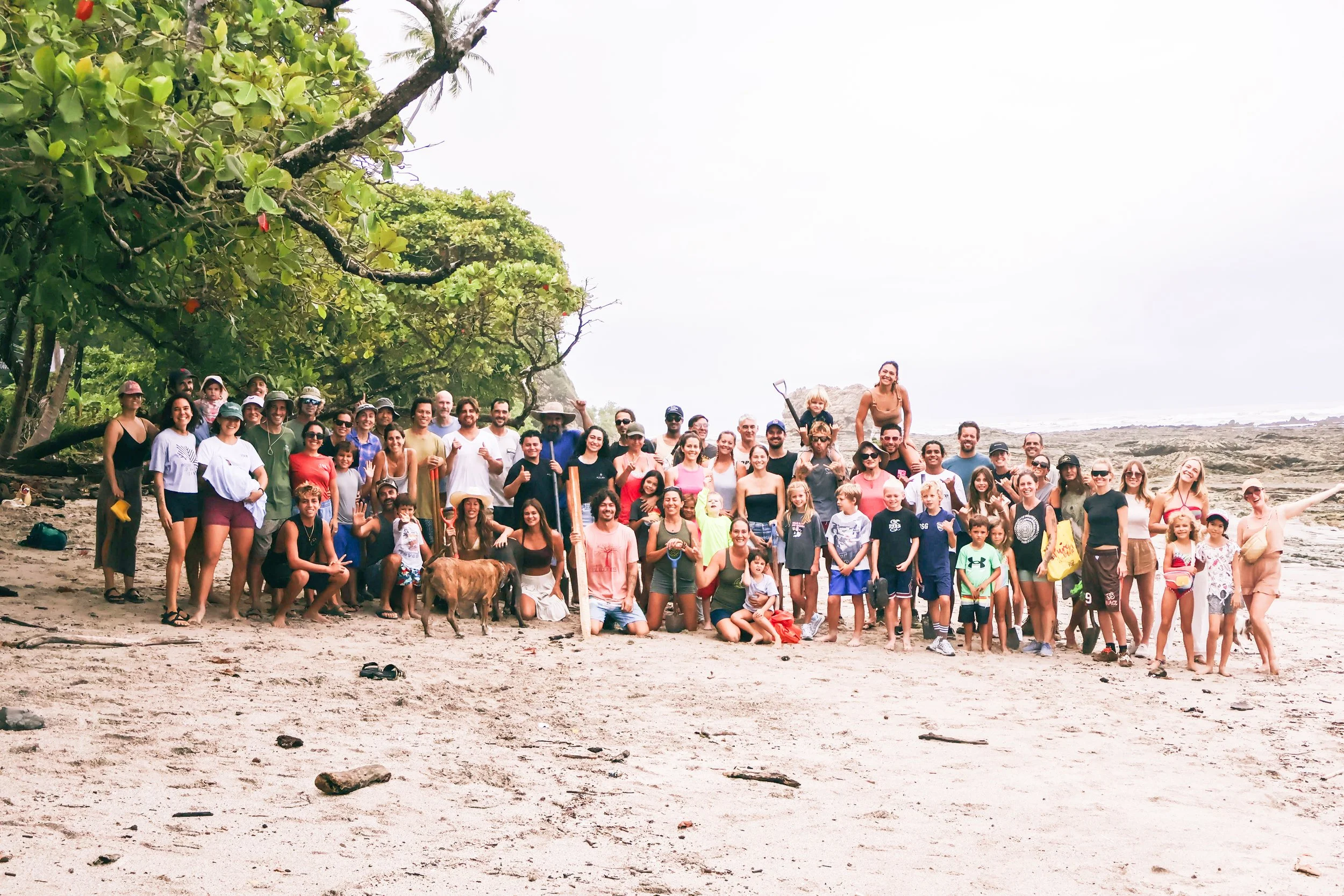 Hermosa Community Group. Large group of people on a beach, with some wearing hats and sunglasses, children and adults smiling and posing for a photo, rocky shoreline and cloudy sky in the background.