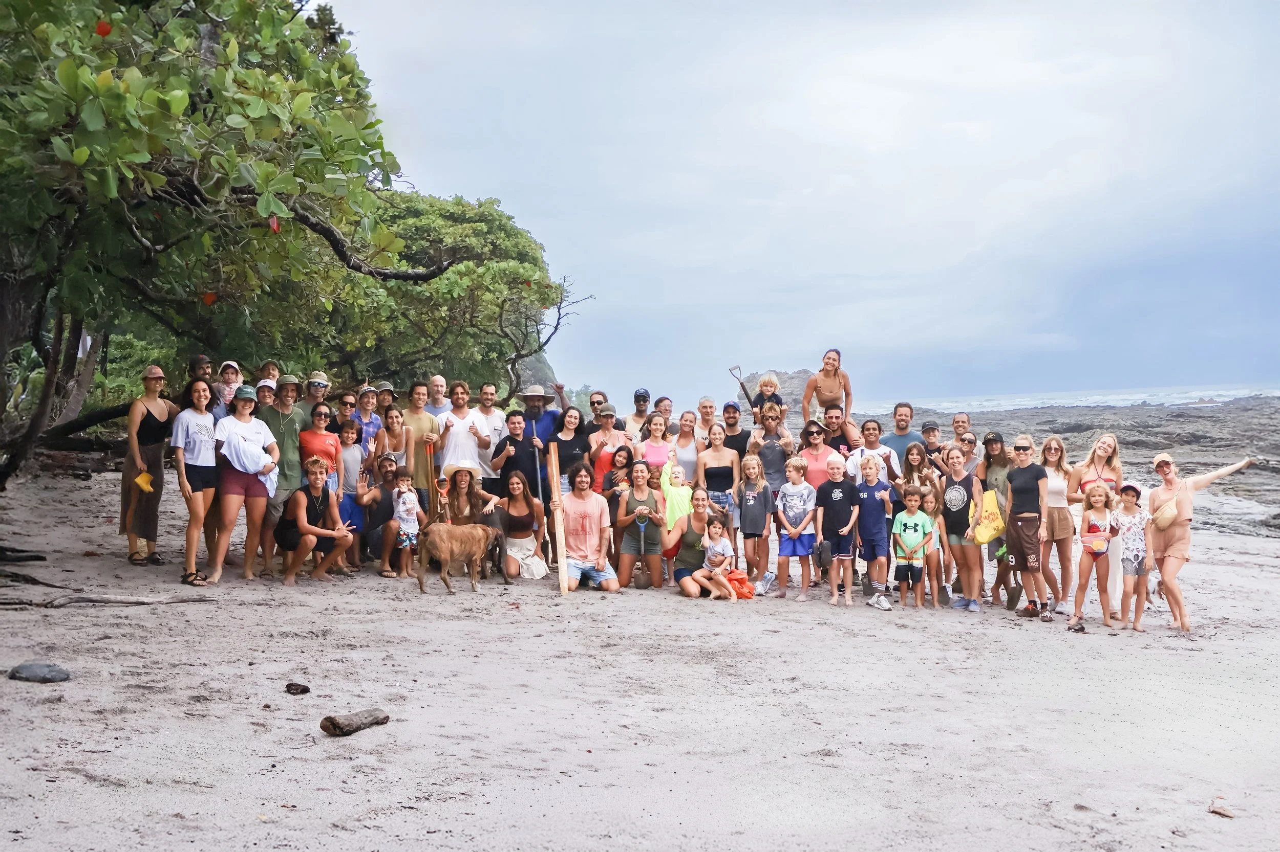 Hermosa Community Group. Large group of people on a beach, with some wearing hats and sunglasses, children and adults smiling and posing for a photo, rocky shoreline and cloudy sky in the background.