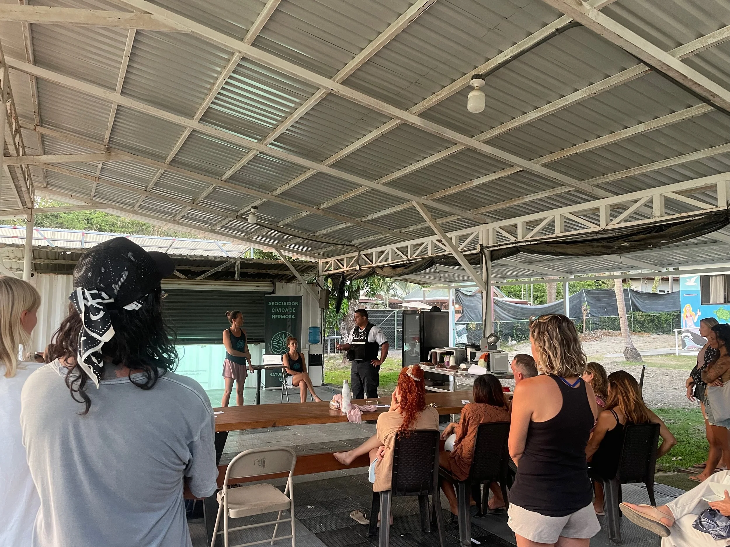 Group of people seated and standing under a covered outdoor area, attending a presentation or meeting, with a woman speaking at the front and a sign that reads 'Asociación Cívica de Hermosa'.
