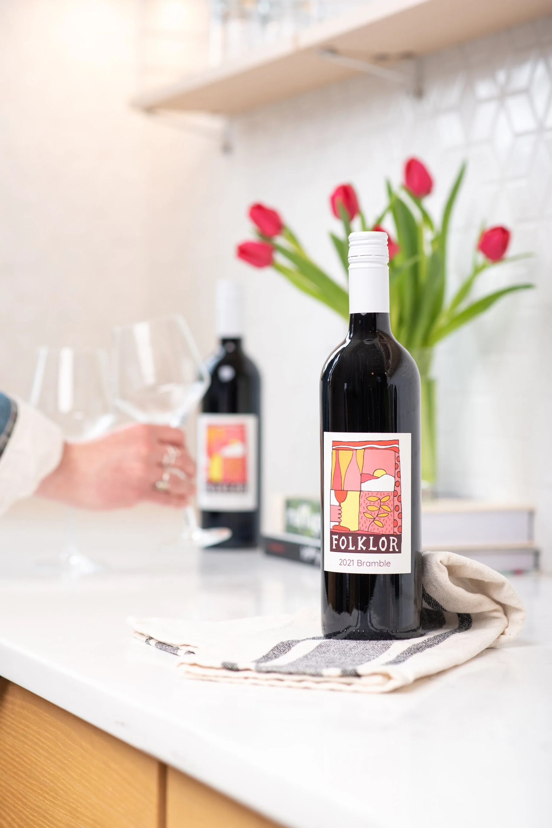 Two Folklore red wine bottles with a woman setting two wine glasses down on the counter. Northern Michigan Marketing Agency