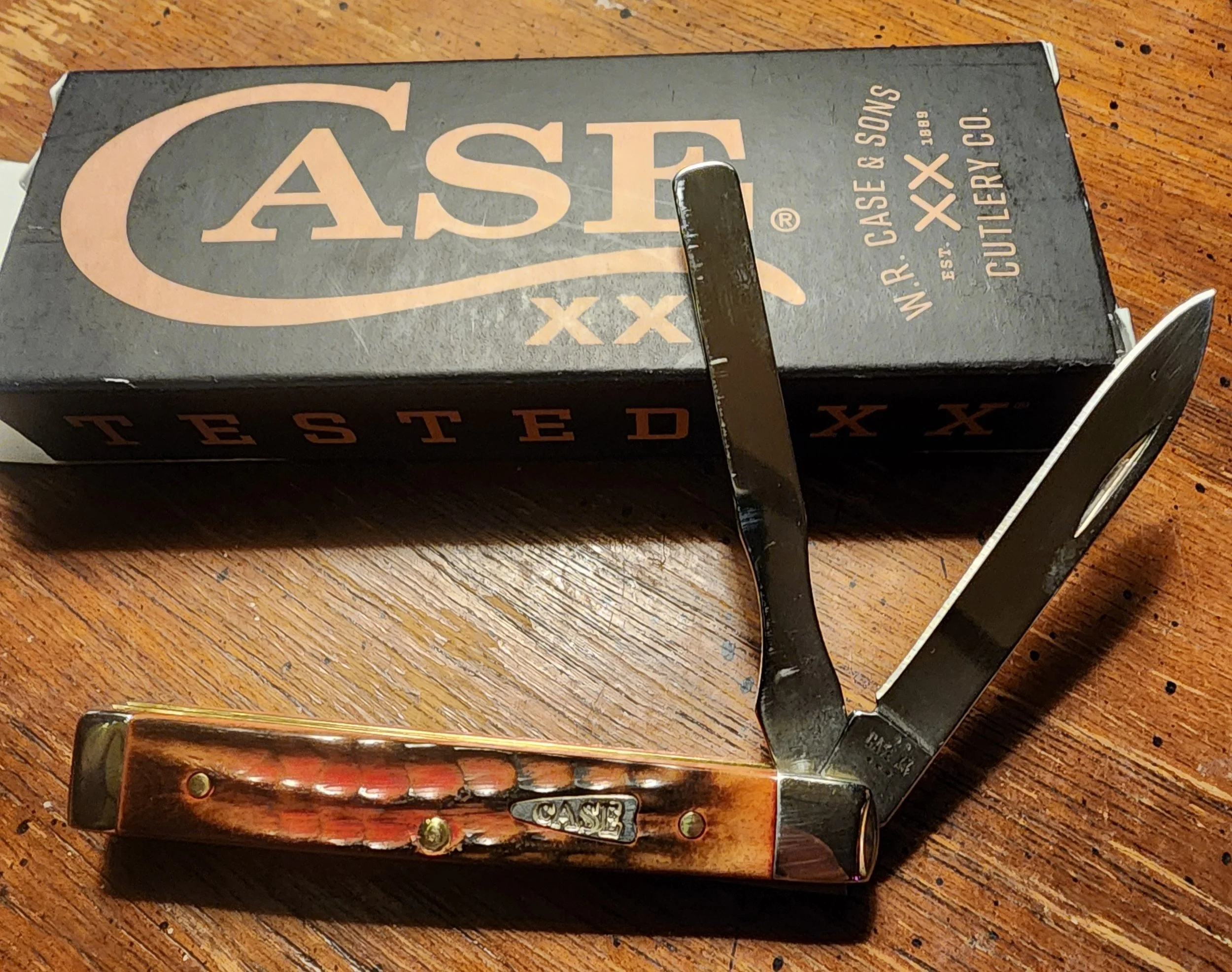 A vintage pocket knife with a wood handle and metal blades, placed next to a black box labeled 'CASE'. The knife is partially open, showing two blades, on a wooden surface.