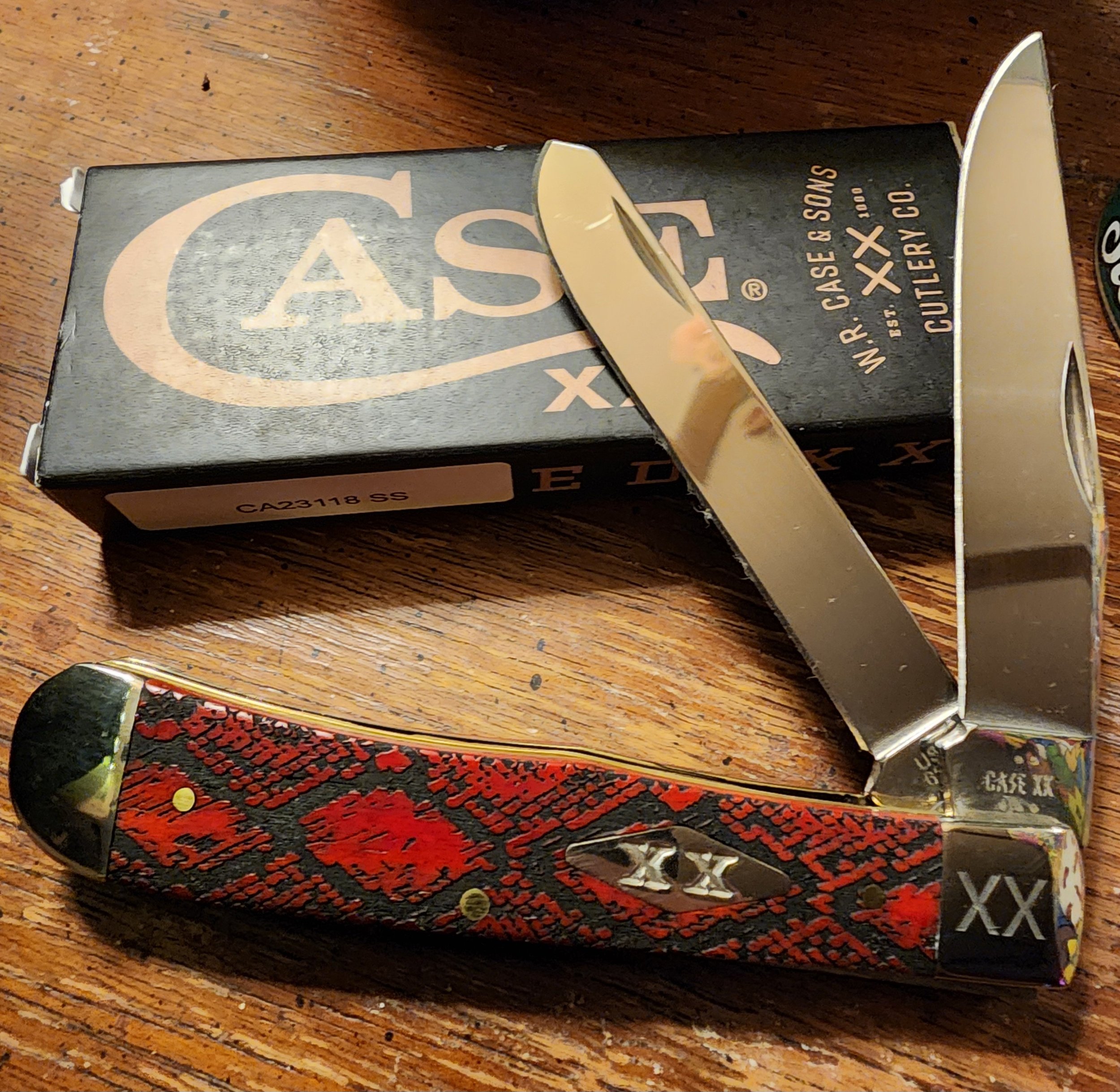 A pocket knife with a red and black snake skin pattern on its handle, placed next to a black box labeled 'CASE XX W.R. CASE & SONS CUTOLEY CO.'