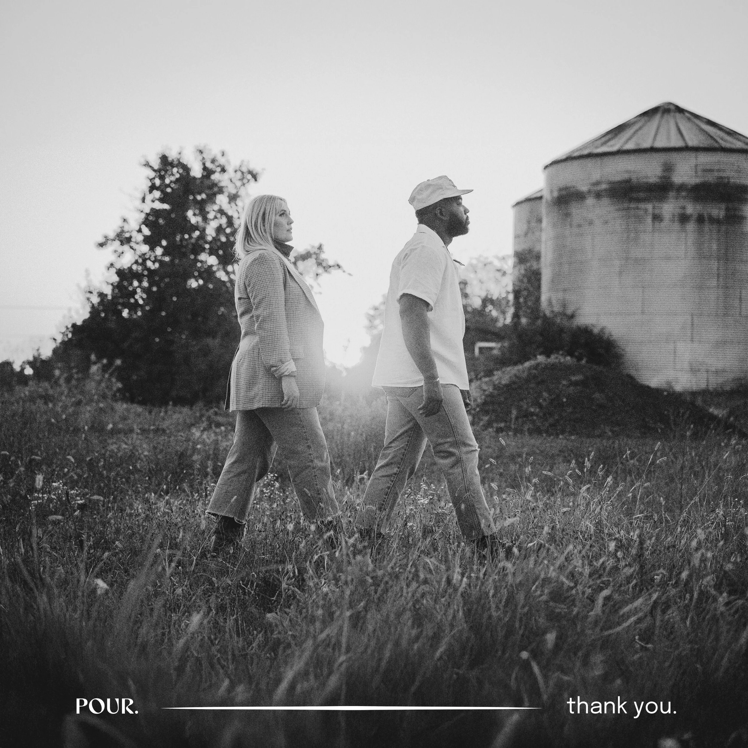Black and white photo of a woman and a man walking through a grassy field with two silos in the background, at sunset.