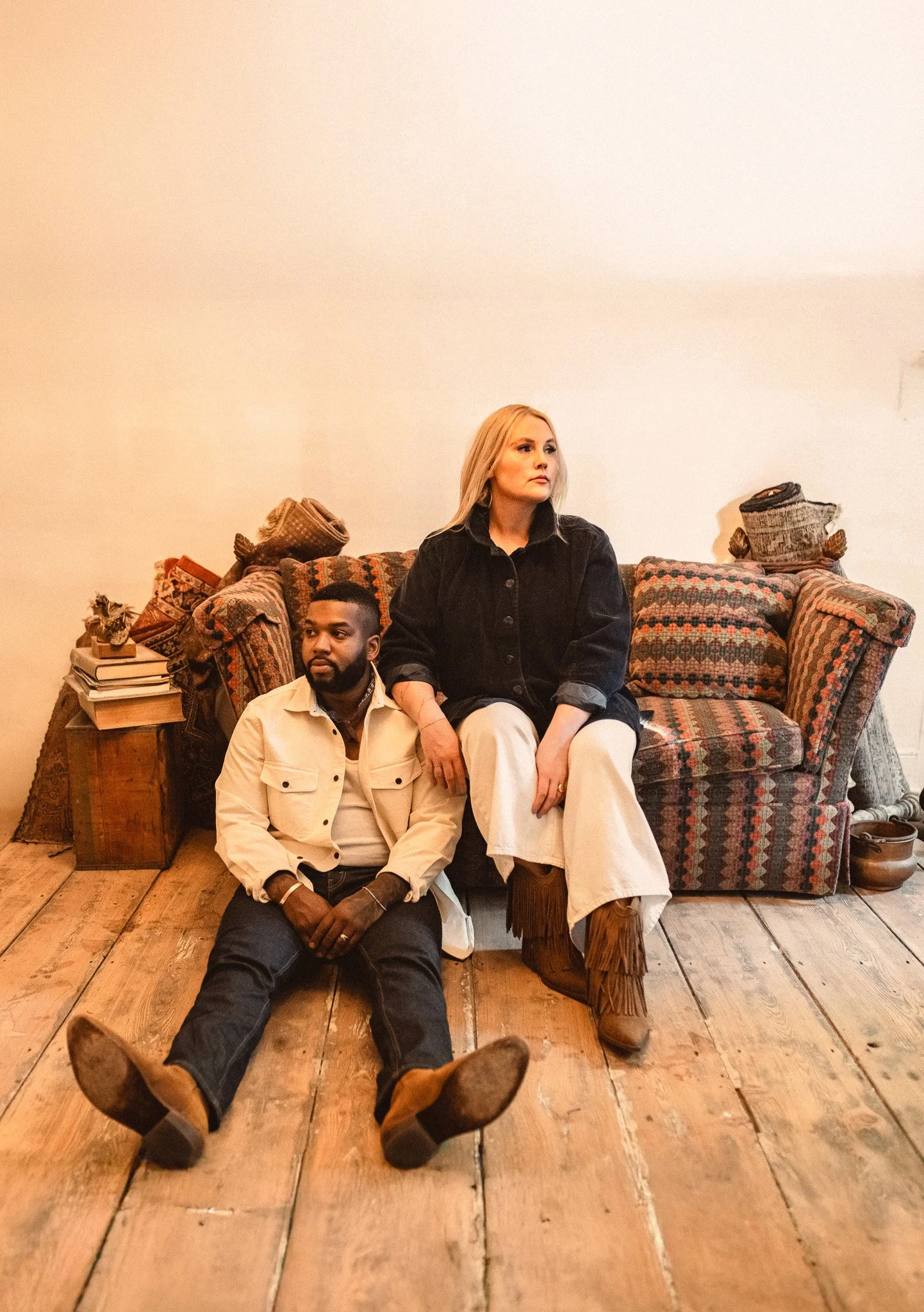 Two people sitting on the floor in front of a vintage sofa. The man is leaning against the sofa, wearing a cream jacket and jeans. The woman is sitting on a wooden block, wearing a black jacket, cream pants, and fringed boots. The room has wooden flo