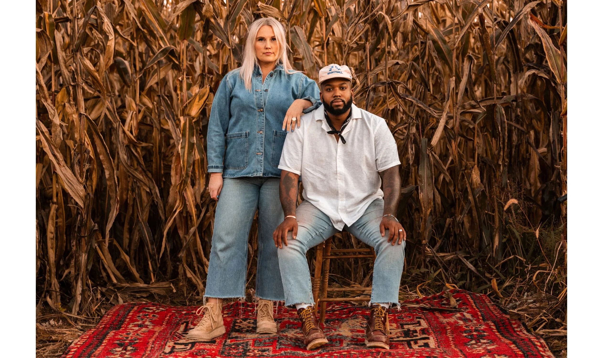 A woman and a man posing together outdoors in a cornfield, standing and sitting on a red patterned rug, with the woman resting her hand on the man's shoulder.
