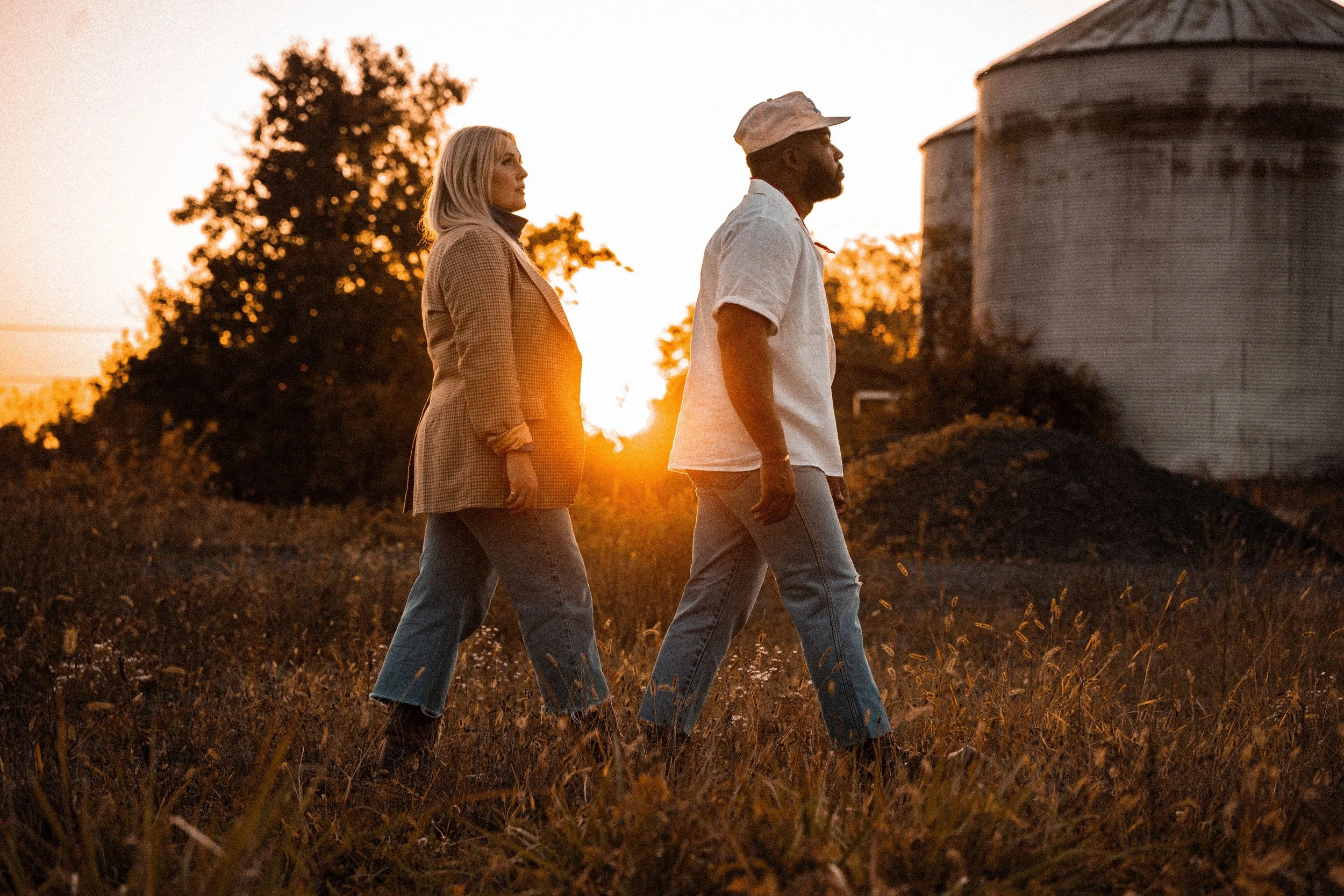 A man and woman walking through a field at sunset, with farm buildings in the background.