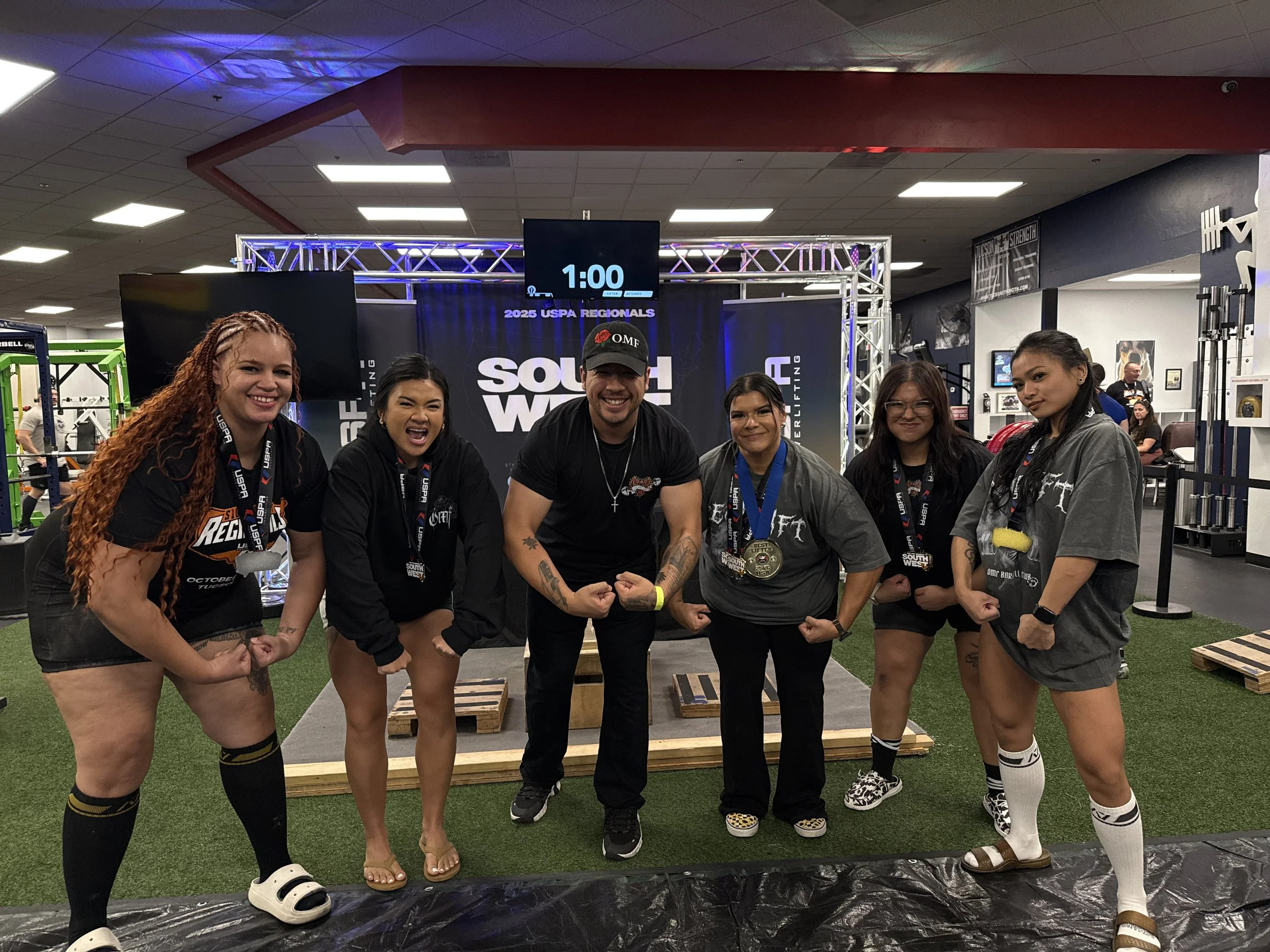 Group of six female powerlifters and one male coach or competitor at a strongwoman competition, posing with medals, flexing muscles, on a stage with equipment and a digital clock in the background.