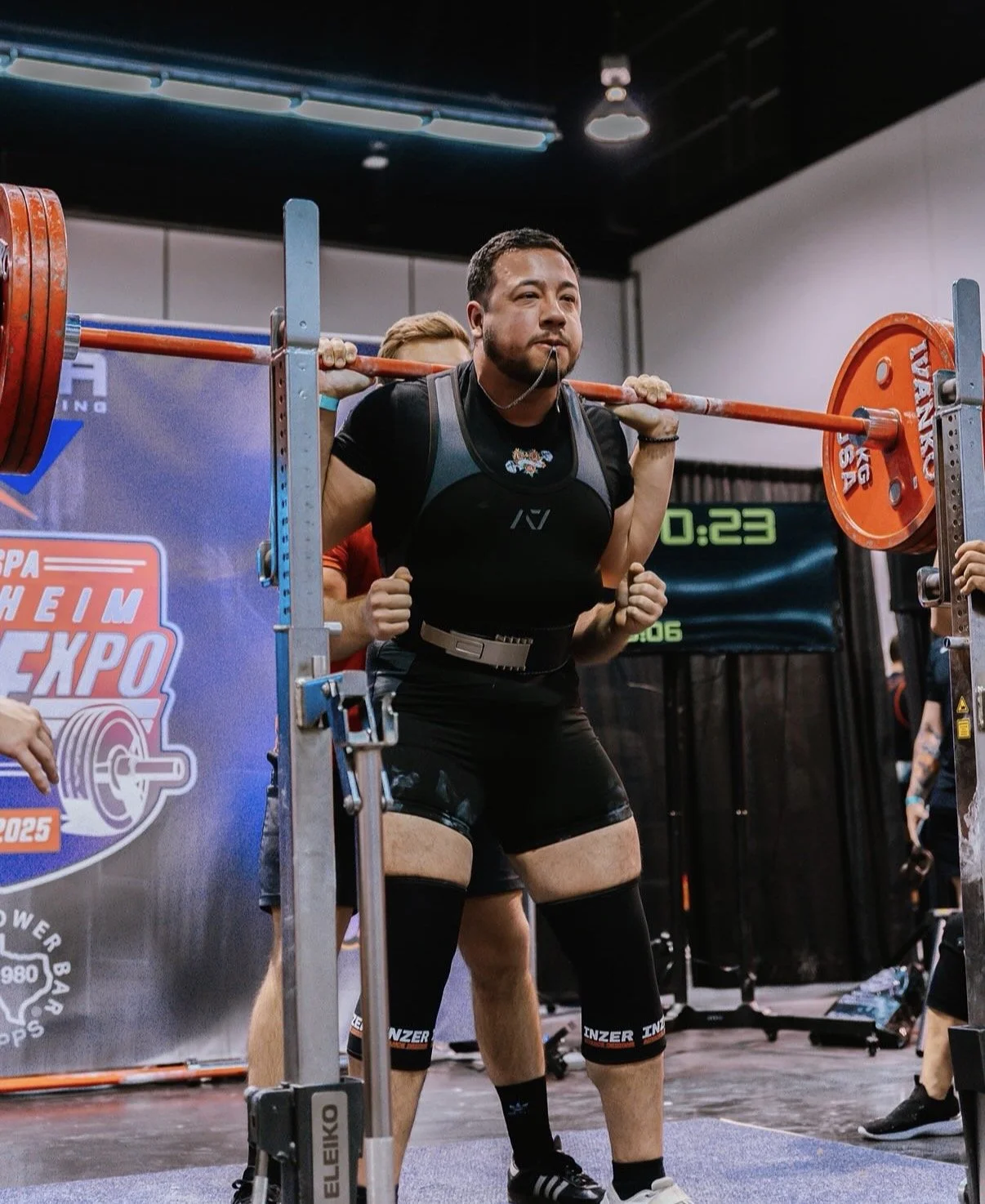 Man lifting a barbell during a powerlifting competition at the Shleim Expo, with a digital timer in the background.