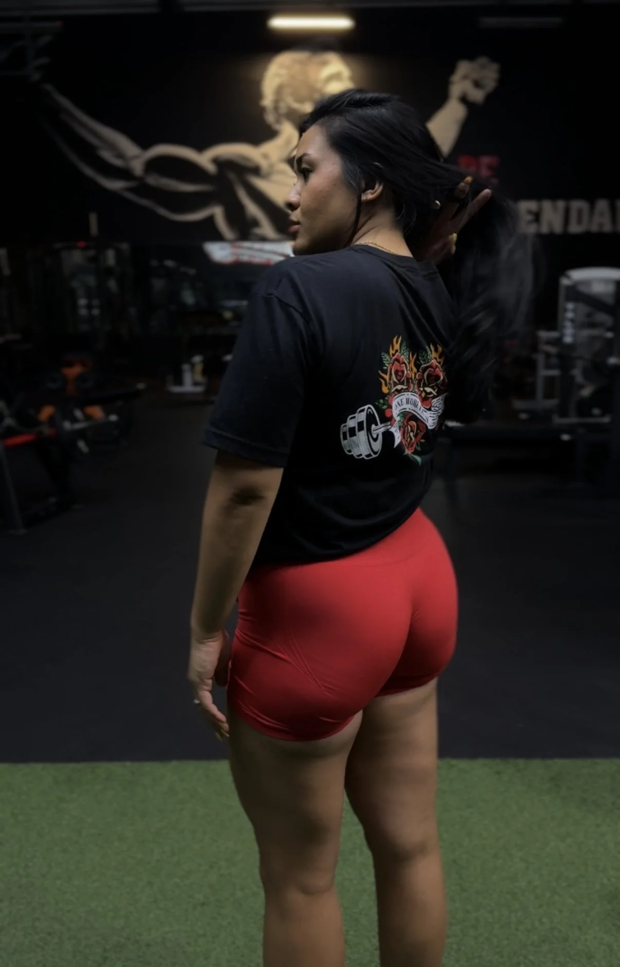 A woman in a gym wearing a black T-shirt with a colorful tattoo design on the back and red athletic shorts, standing on a green mat with gym equipment and a wall mural in the background.