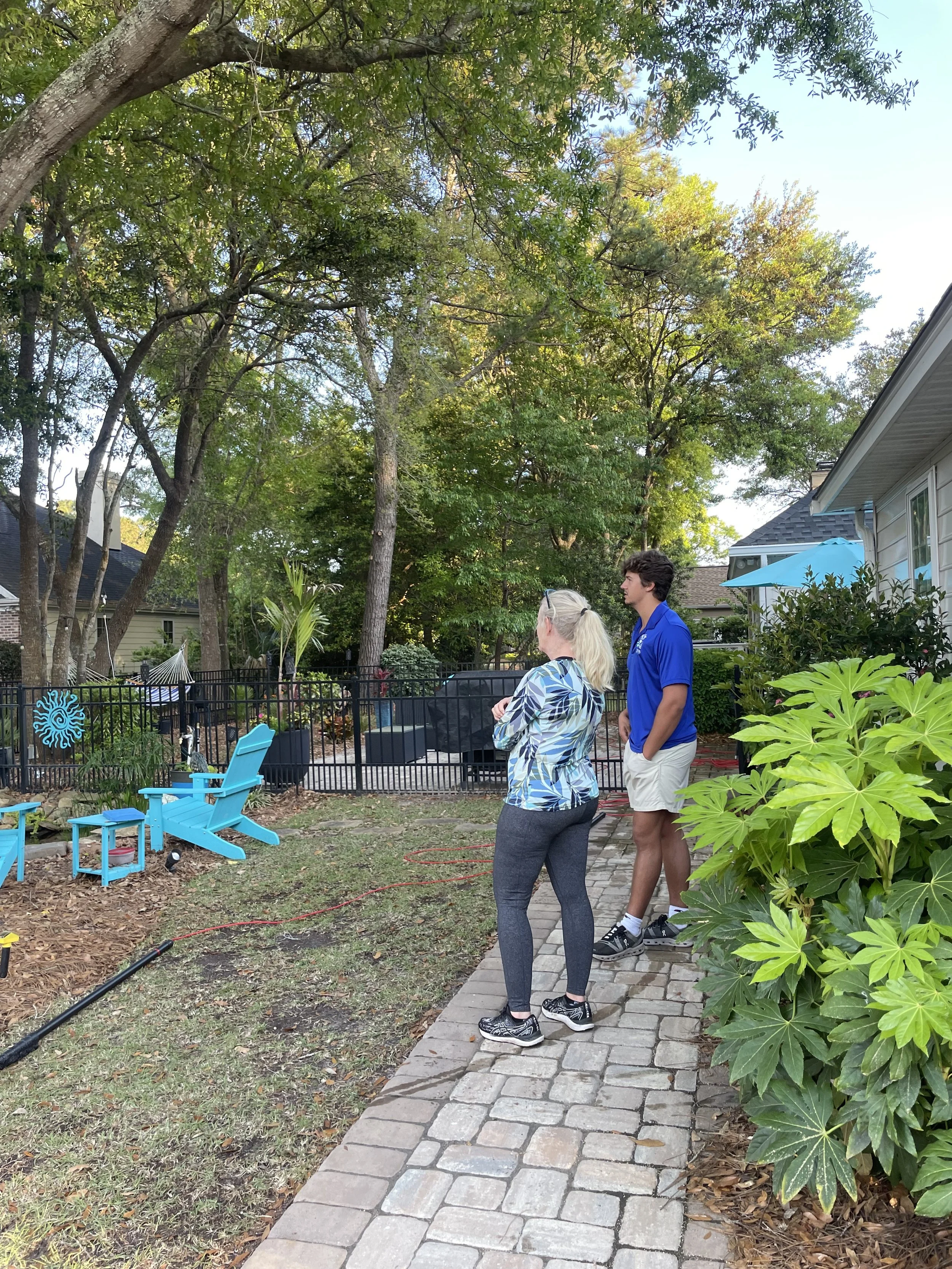 Two people, a woman with blonde hair in a ponytail and a man with dark hair, are standing on a paved walkway in a backyard, talking and looking towards a black fence. The backyard has a green lawn, trees, outdoor furniture, and a small pool area behind the fence. The scene is bright with clear skies.
