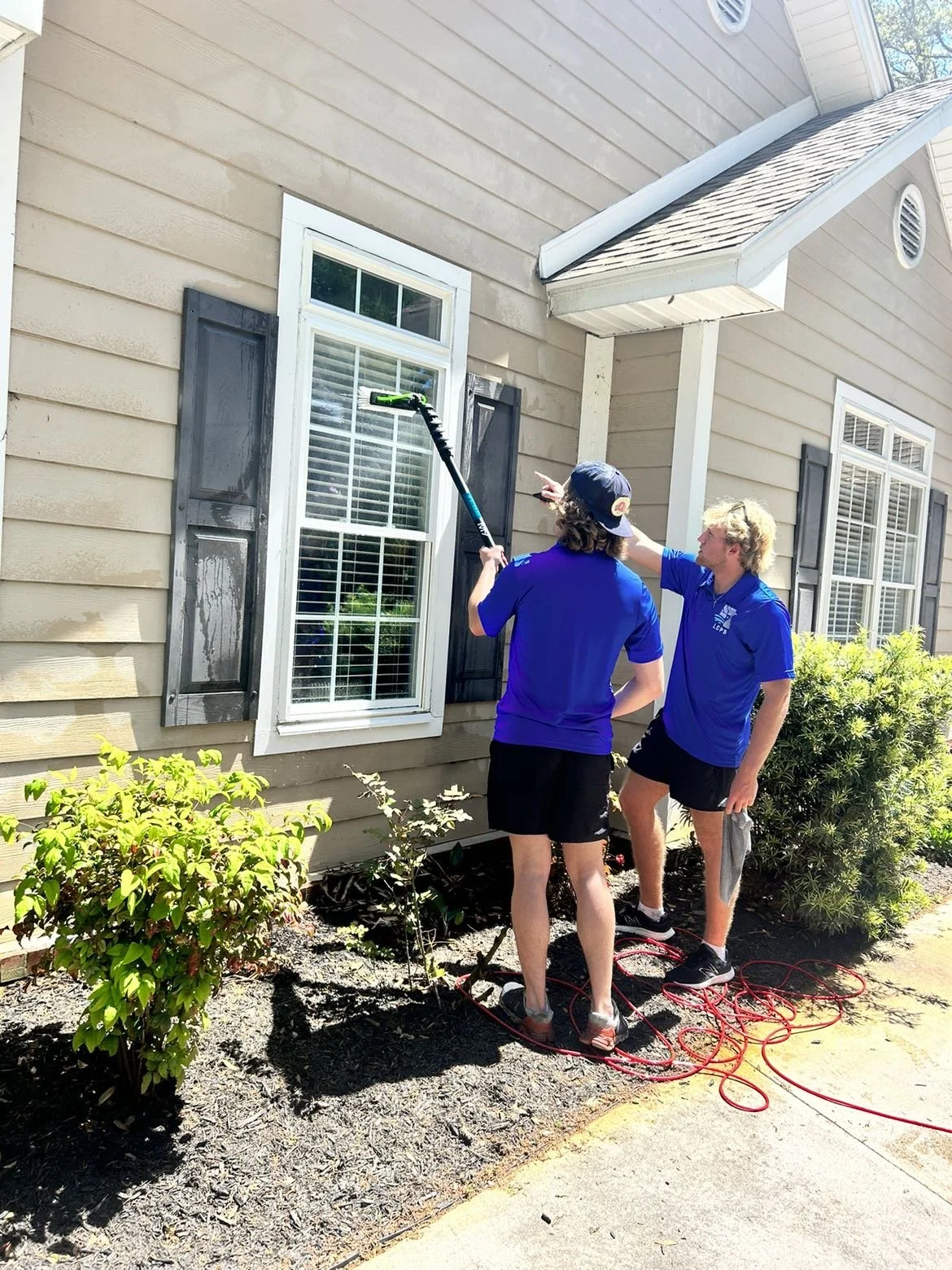 Two young men pressure washing the exterior wall of a house with a window and black shutters, standing on a concrete sidewalk with gardening bushes nearby.