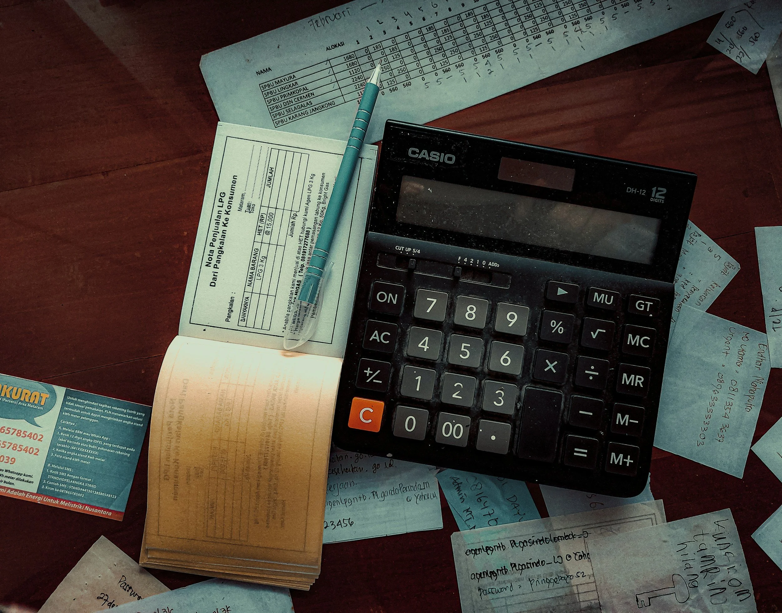 A cluttered wooden table with scattered documents, a calculator, a blue pen, and various handwritten notes and bills.