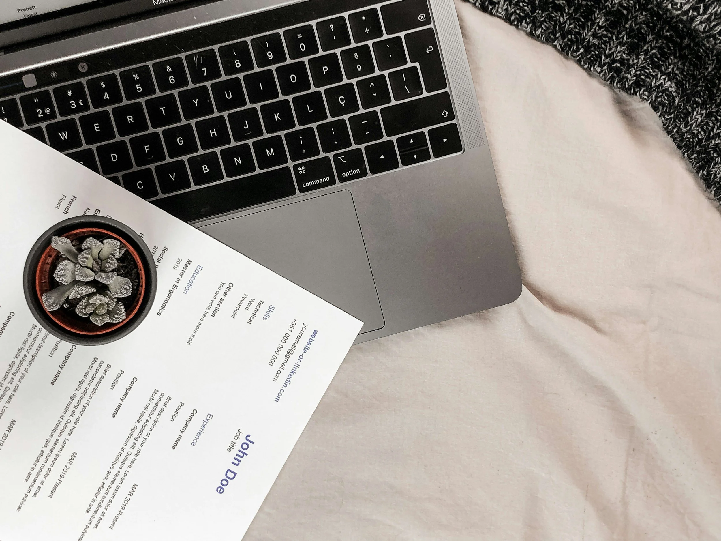 A silver MacBook laptop on a bed with a white sheet and a gray knitted blanket, partially covering a printed document with a small potted cactus plant on top.
