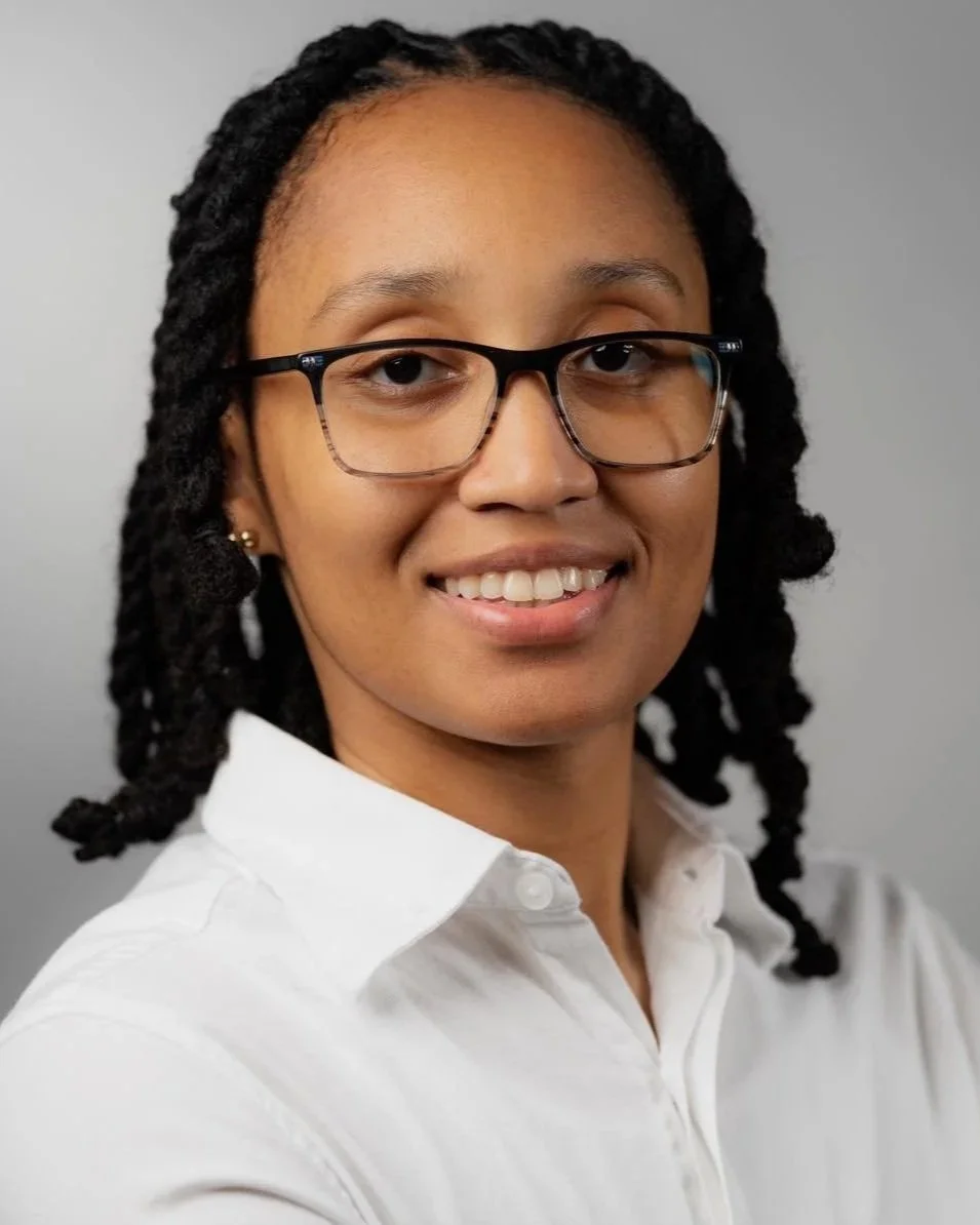 Close-up portrait of a woman with glasses, dark hair styled in twists, wearing a white collared shirt, smiling against a plain background.