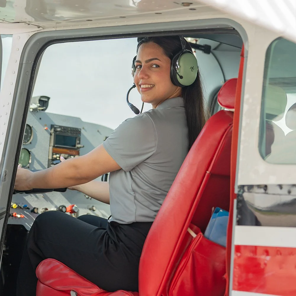 A woman sitting in the cockpit of an airplane, wearing a headset, with a smile, looking back over her shoulder.