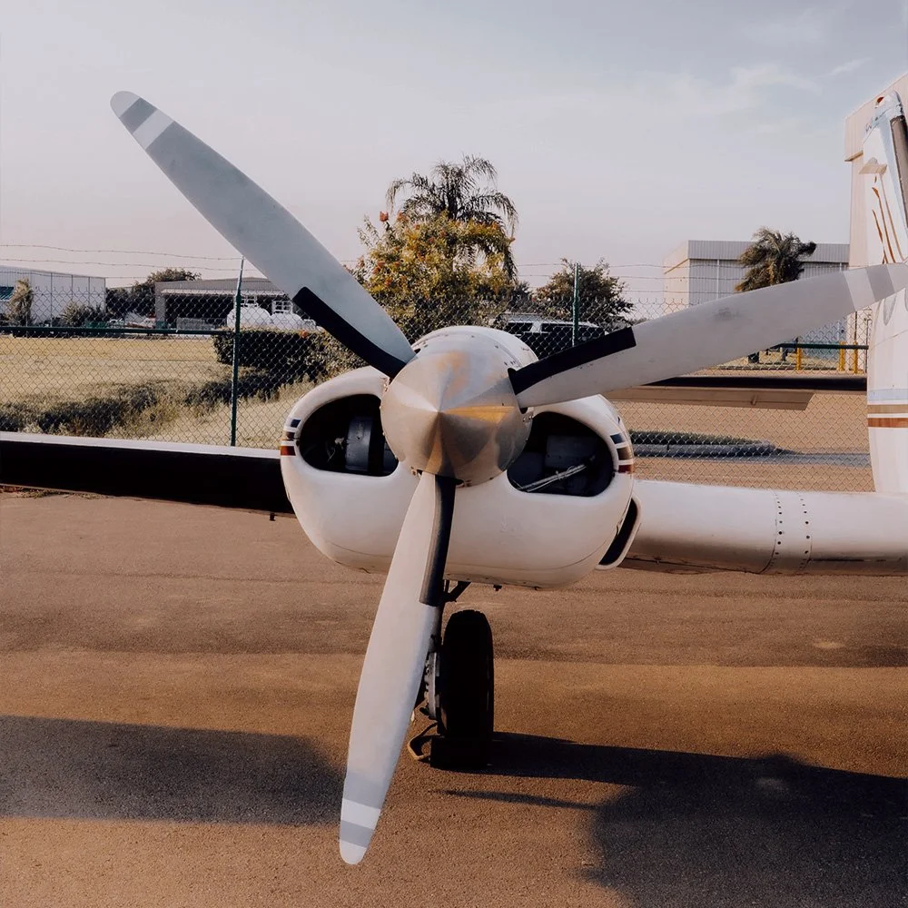 Front view of a single-engine propeller airplane on the tarmac with a chain-link fence, trees, and buildings in the background.