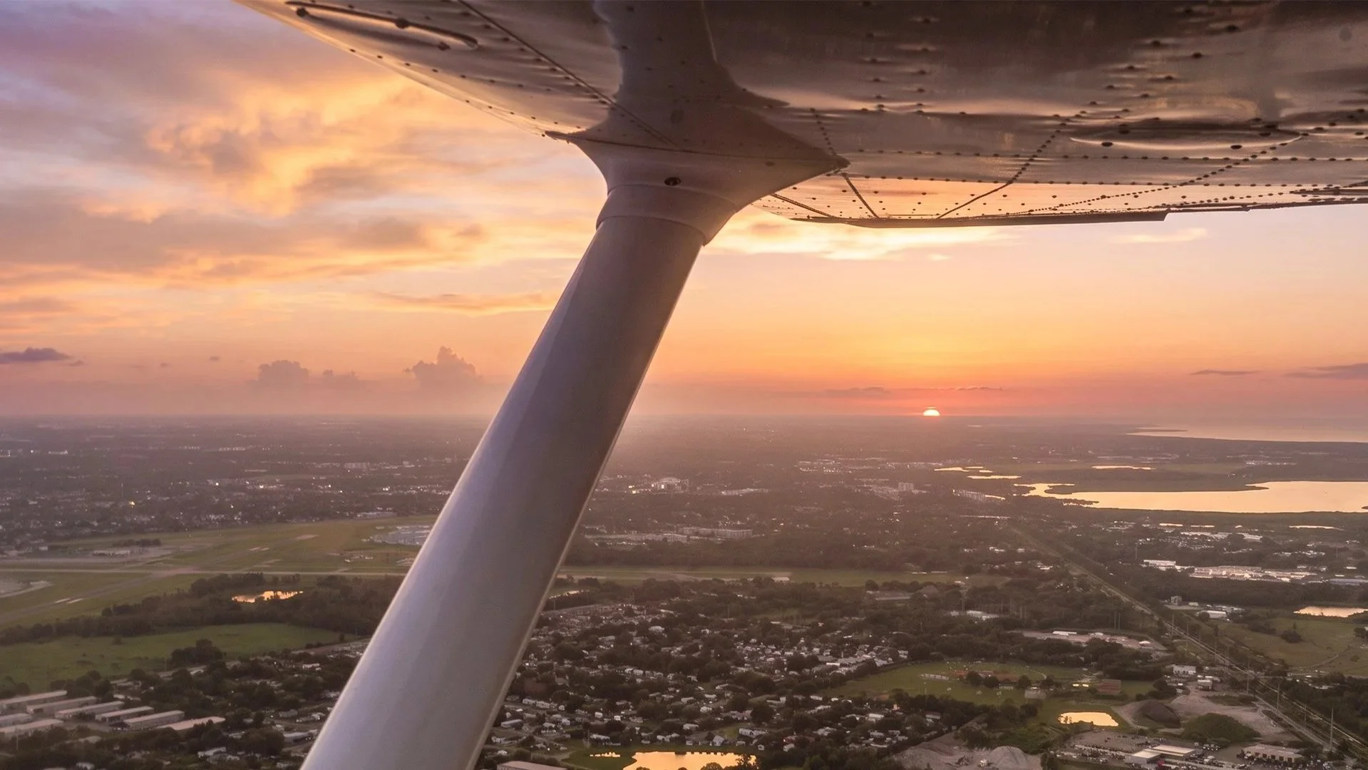 View from a small aircraft window showing a sunset over a landscape with water bodies, fields, and buildings, with the aircraft's wing strut in the foreground.