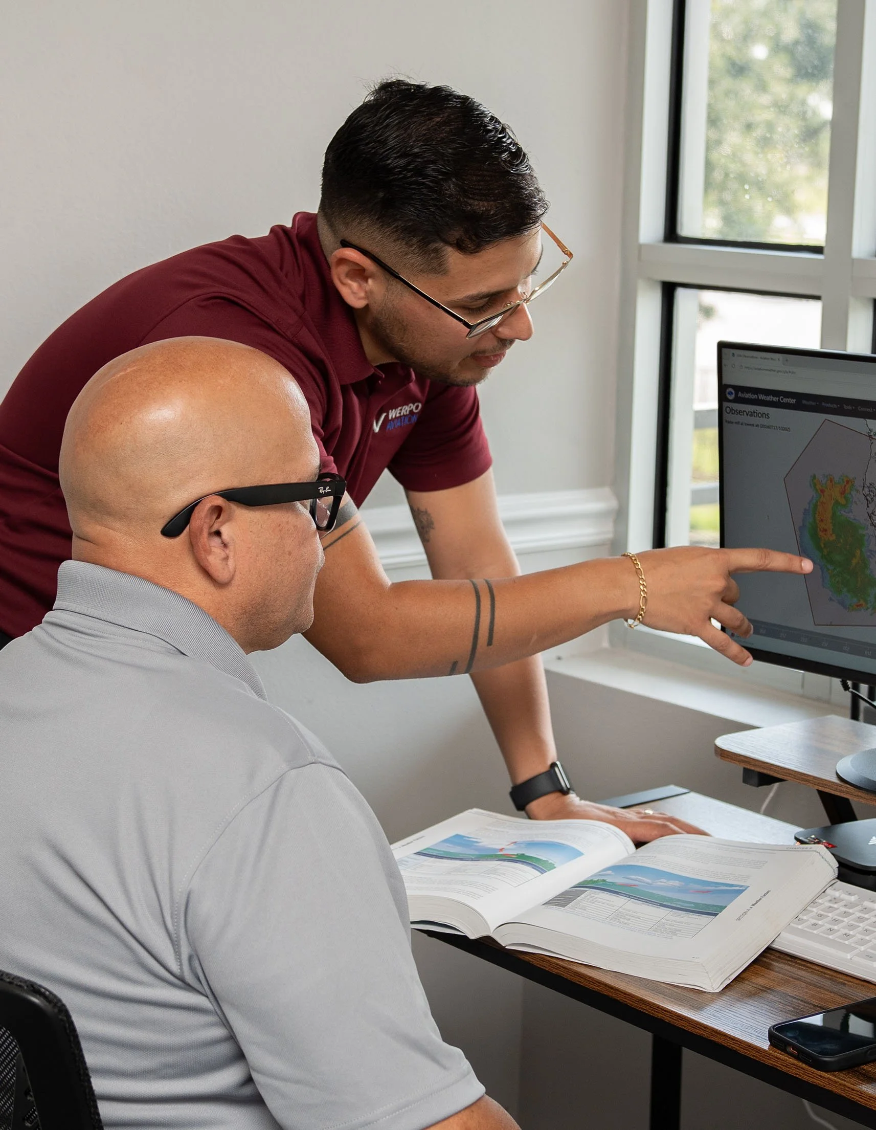 Two men studying weather data at a desk, one pointing at a computer screen showing weather observations, with open weather charts and a window in the background.