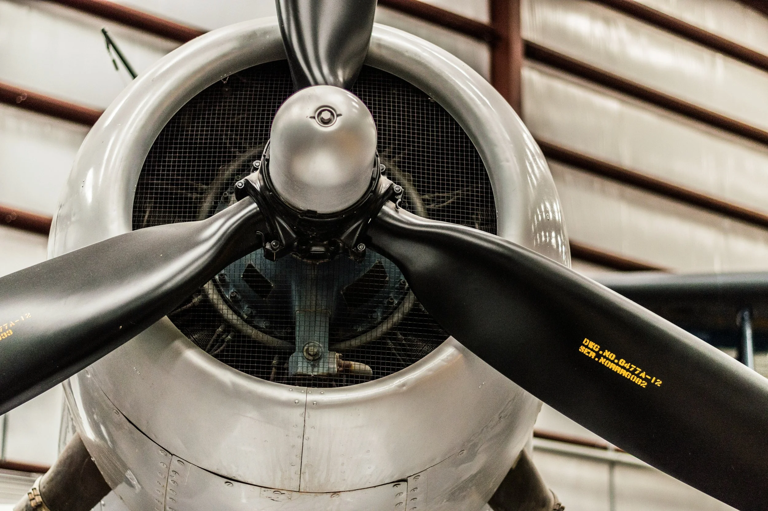 Close-up of the front of an aircraft engine with three black blades, inside a hangar with wooden beams.