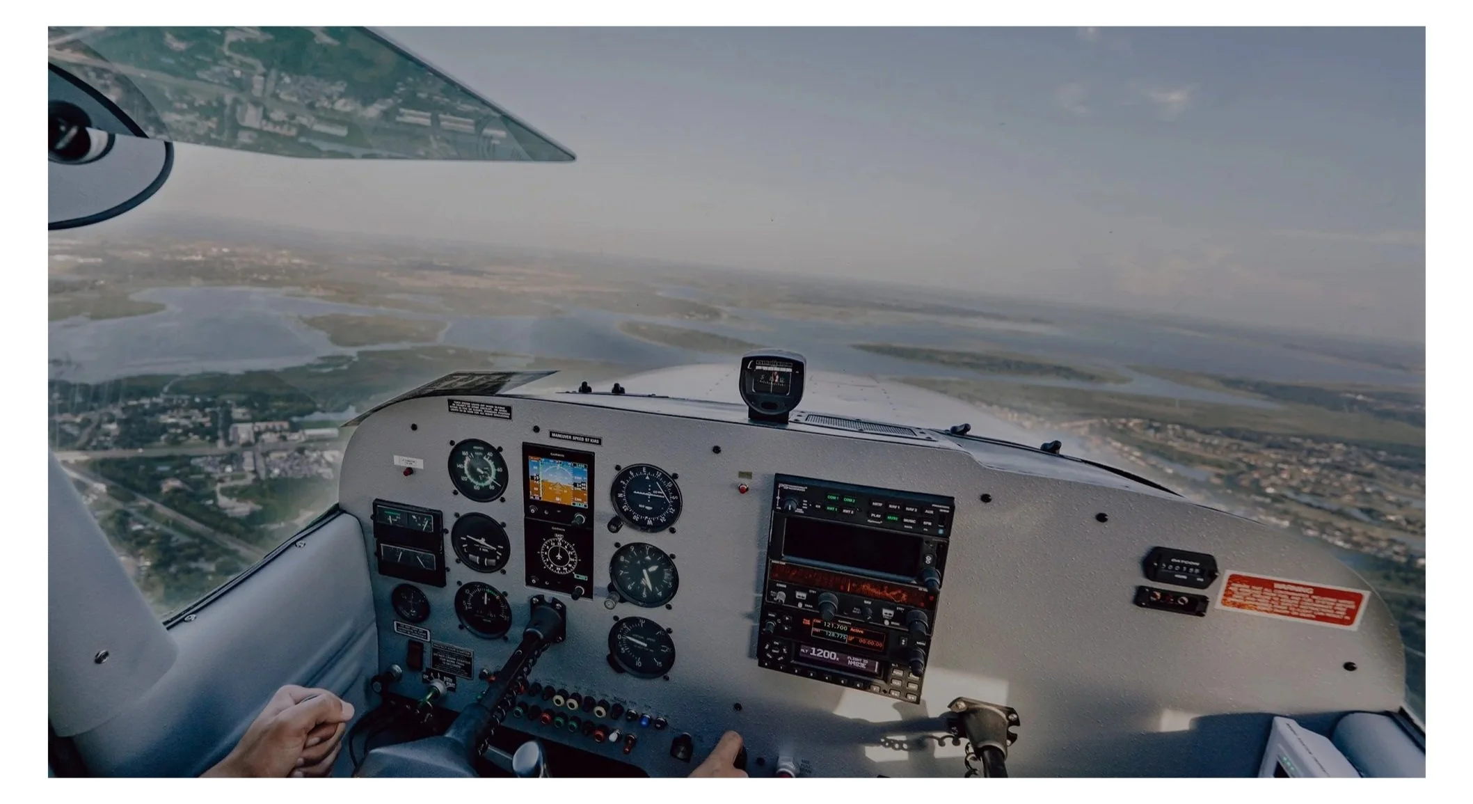 View from the cockpit of a small airplane flying over a landscape with water bodies, land, and a city in the distance.