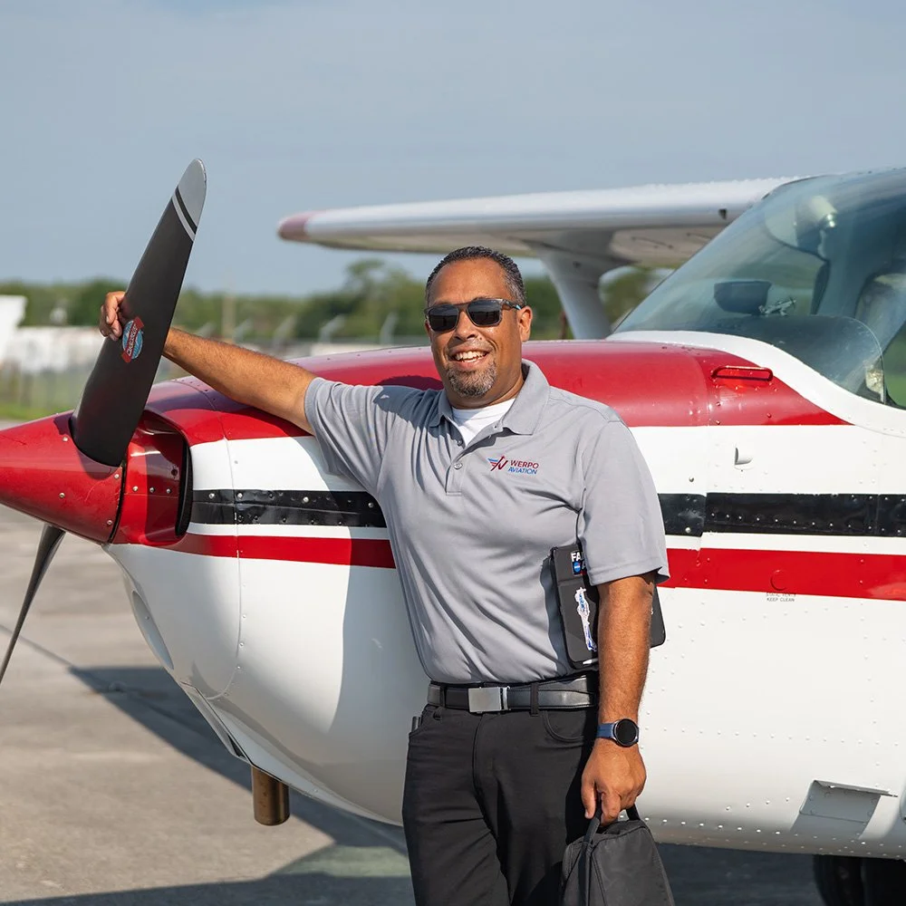 A man in sunglasses smiling and leaning on a small red and white airplane on the tarmac.