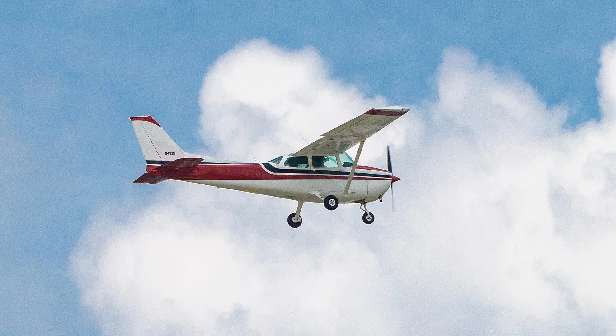 Small white and red propeller airplane flying in a blue sky with scattered white clouds.