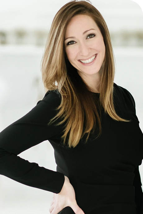 A woman with long, light brown hair smiling and looking at the camera, wearing a black top, standing indoors with a blurred background.
