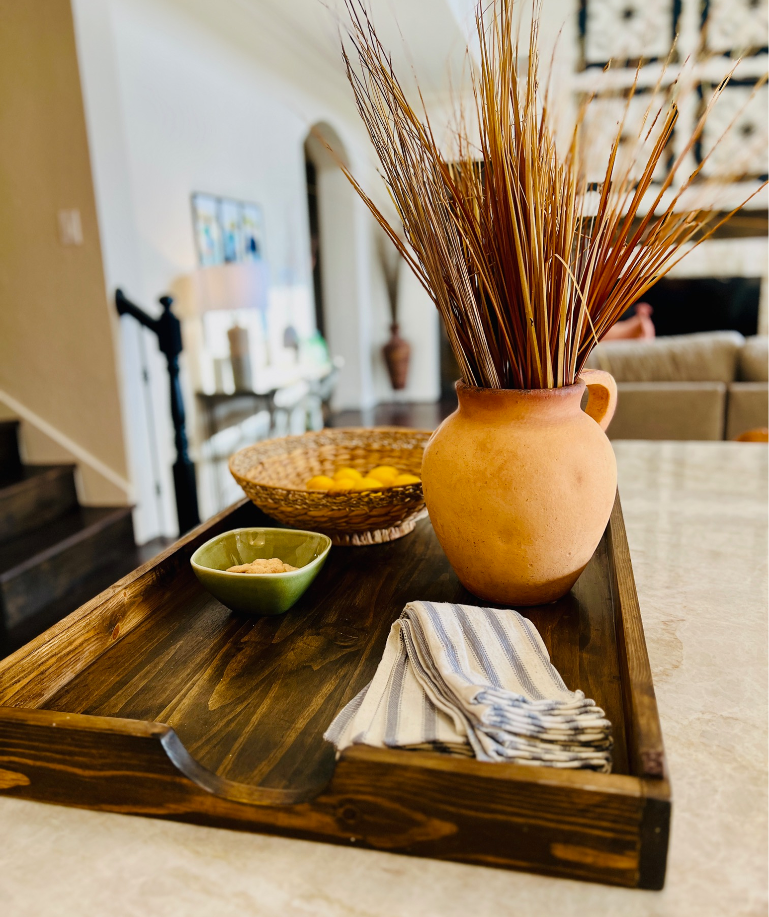 A wooden tray on a kitchen island with a large terracotta vase filled with dried grass, a green bowl with snack food, a woven basket with lemons, and a folded striped cloth napkin.