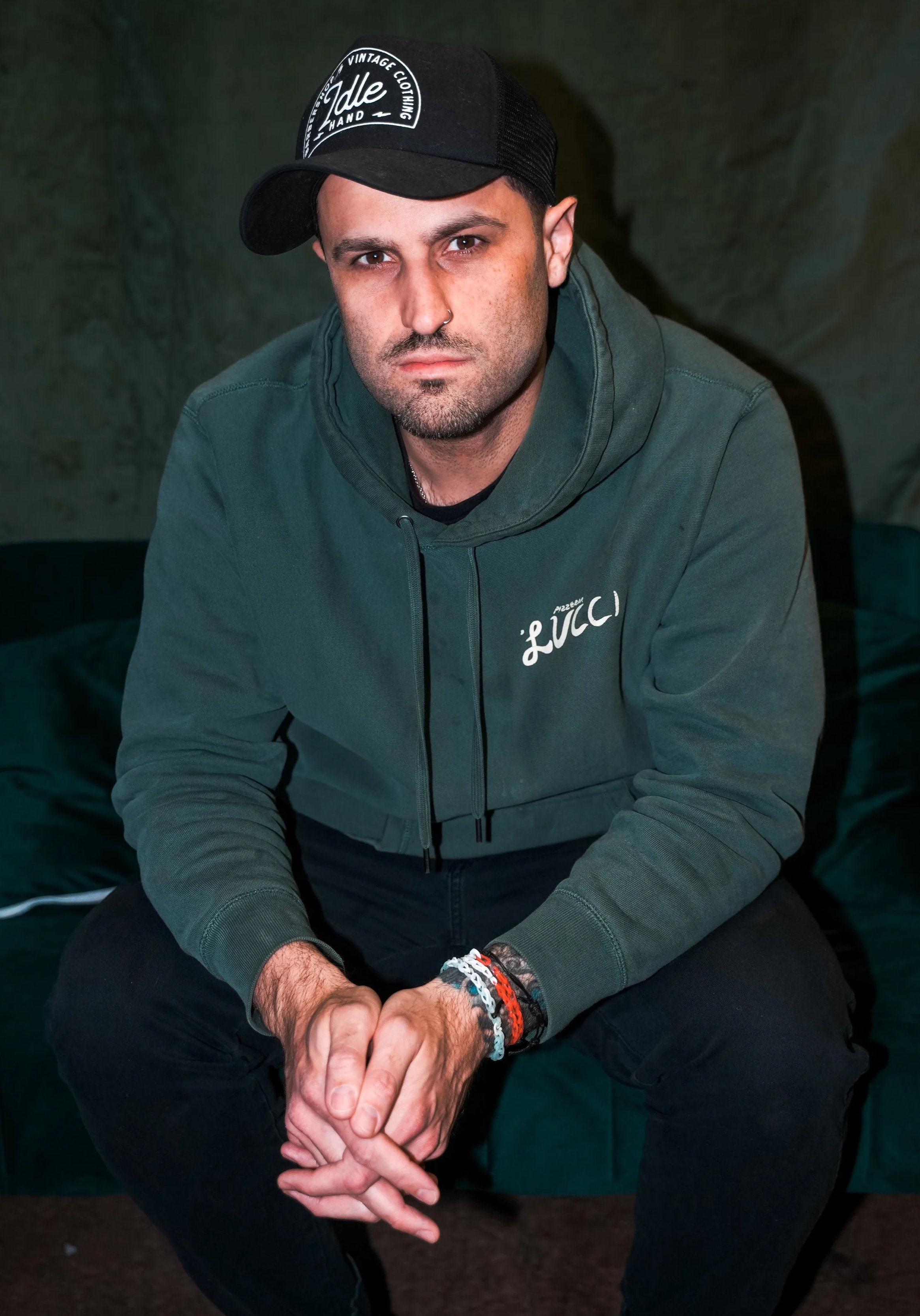 A man wearing a black cap, green hoodie with "Lucci" written on it, sitting on a green sofa with his hands clasped.