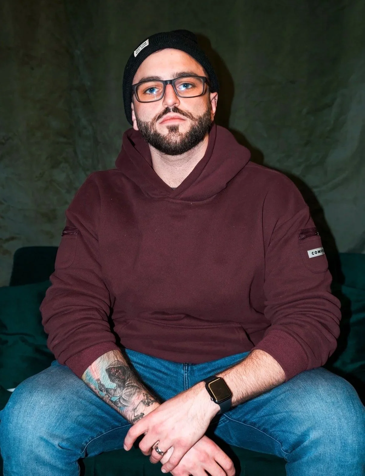 A man with glasses and a beard, wearing a black beanie and a maroon hoodie, sitting with his hands clasped, in front of a dark green background.
