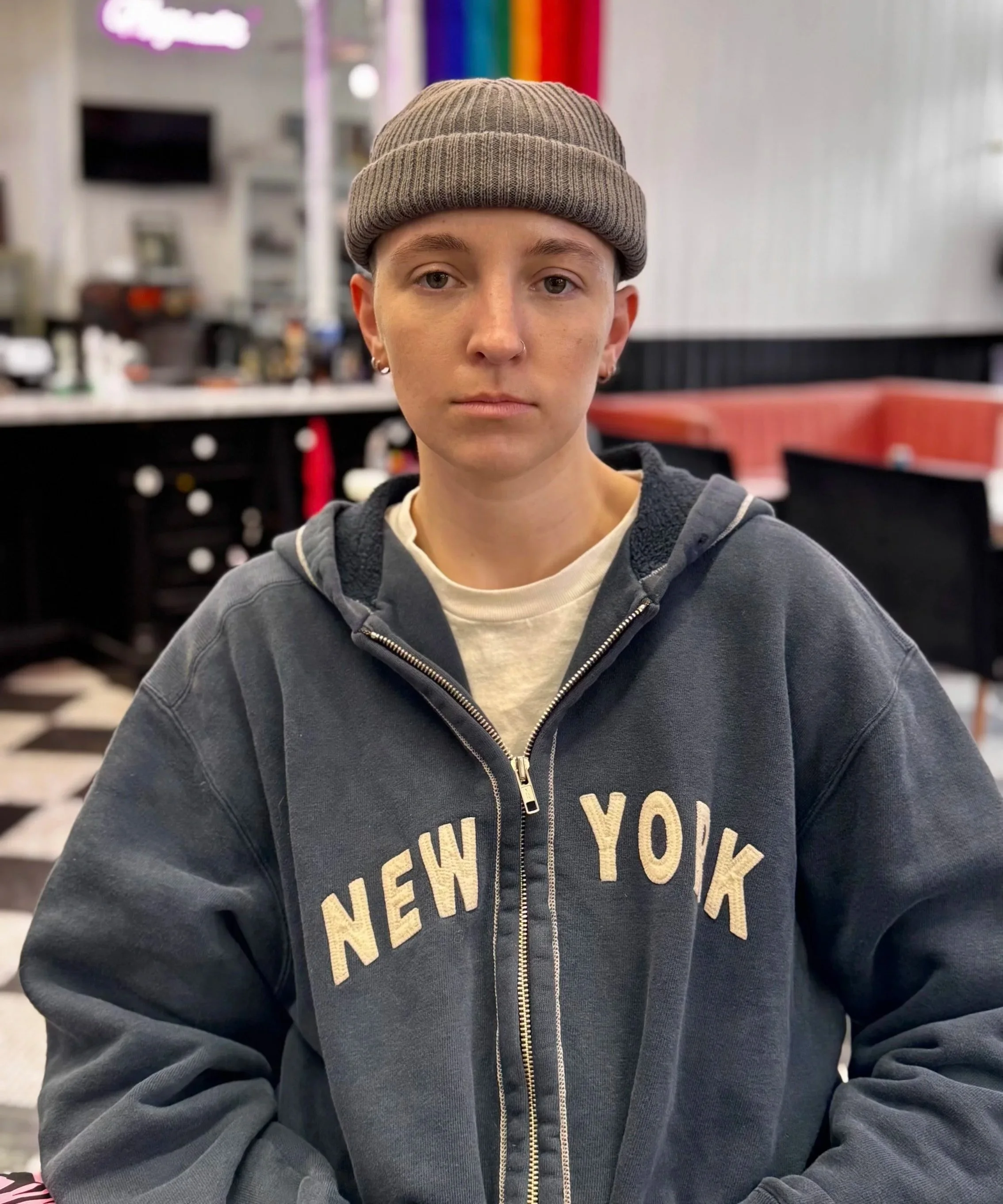 A young person wearing a dark gray hoodie with 'New York' written on it, a beige beanie, and with a neutral expression, sitting inside a cafe with a colorful background.