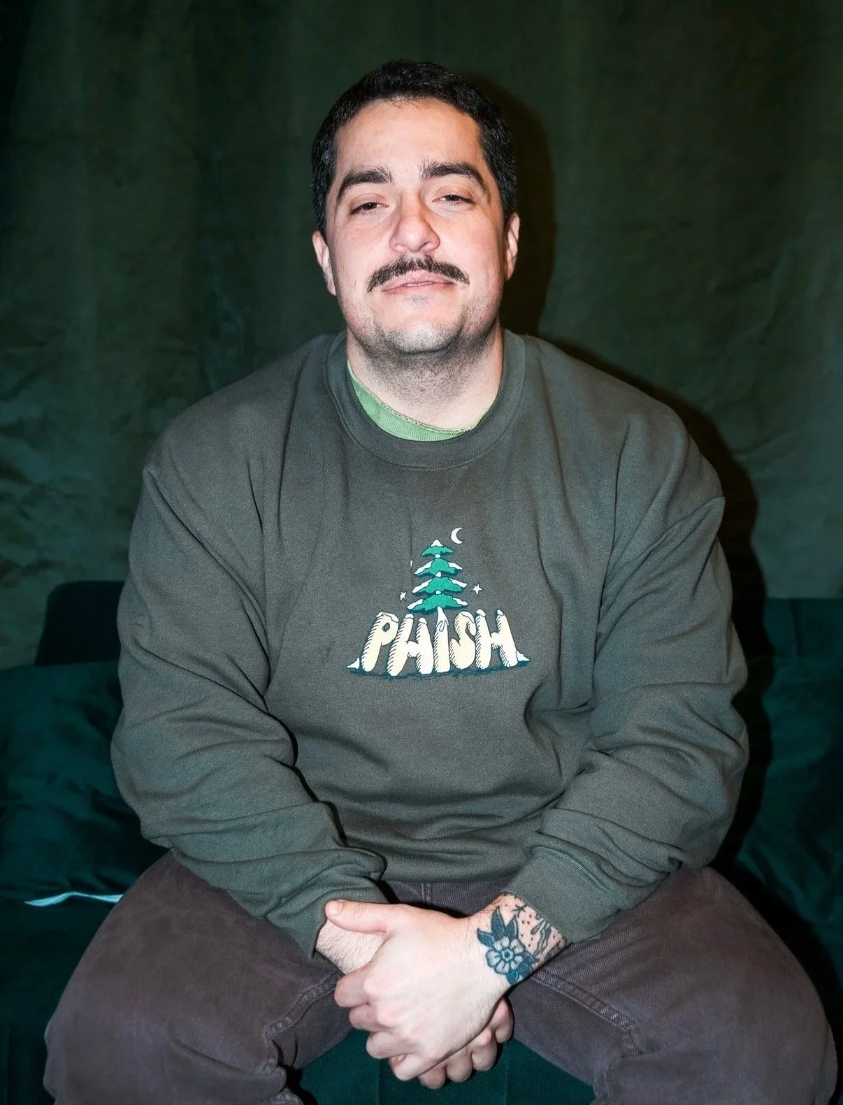 A man with dark hair, mustache, and light skin is sitting on a couch in front of a dark background, wearing a green sweatshirt with a graphic of a pine tree, moon, and stars, and the word "PLUSH" printed on it. He has a tattoo on his left forearm.