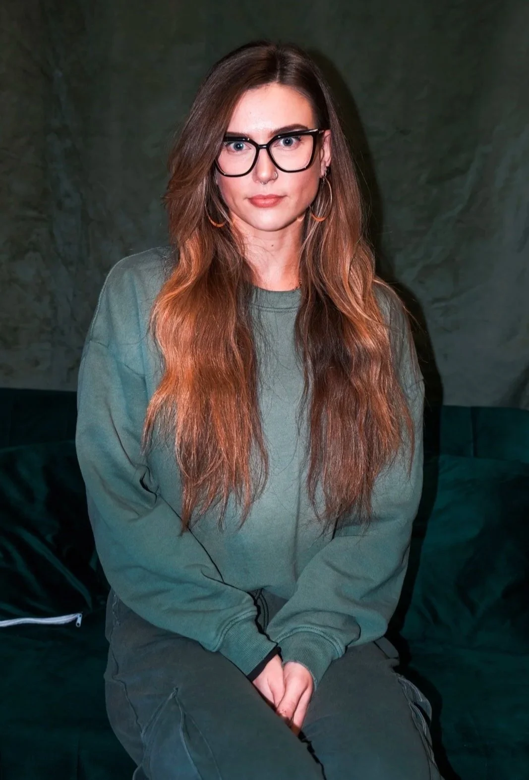 A woman with long reddish-brown hair, wearing glasses, a septum piercing, hoop earrings, a green sweatshirt, and dark pants, sitting on a dark green sofa in front of a textured wall.