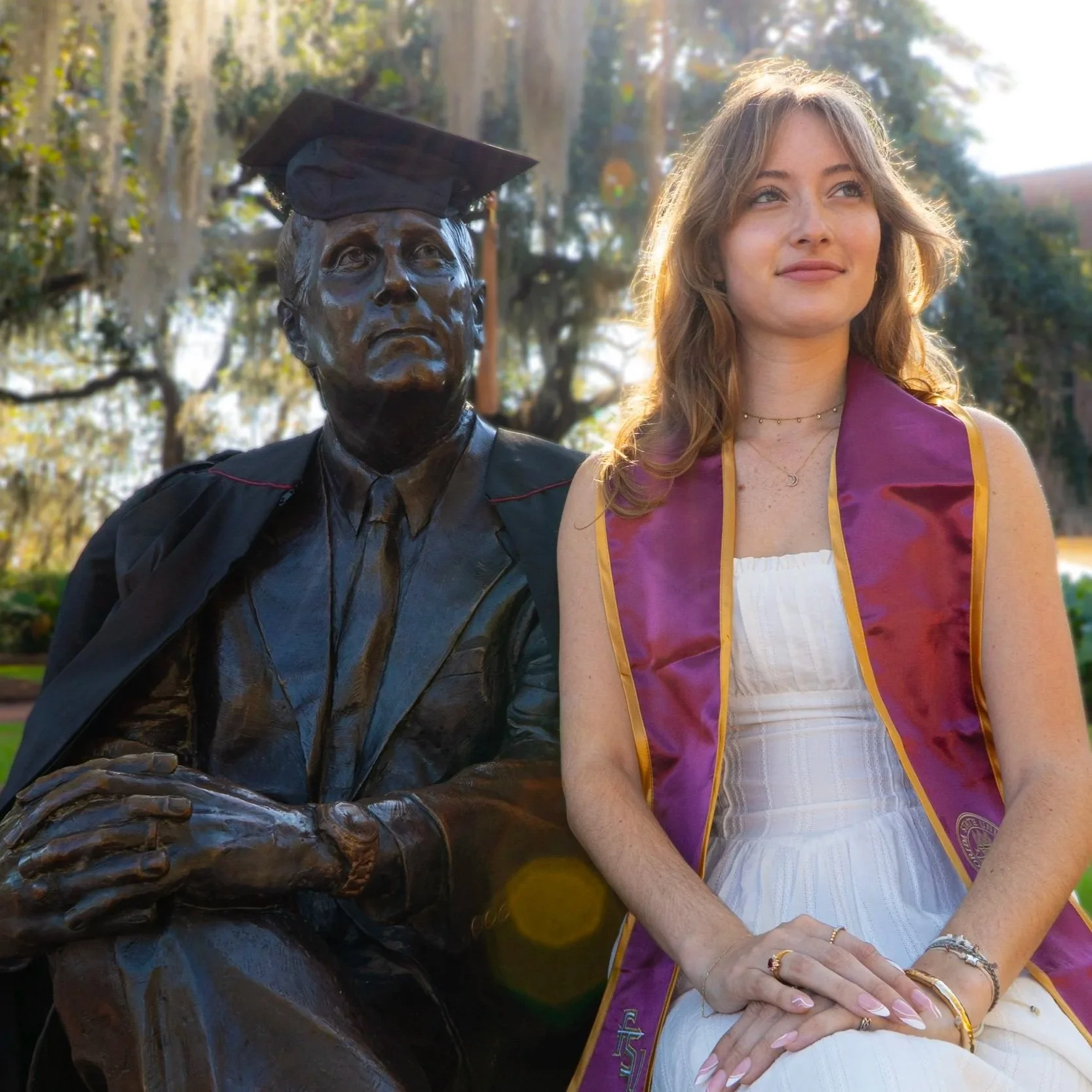 A young woman in a white dress and graduation stole sitting next to a bronze statue of a man wearing a cap, outdoors with trees in the background.