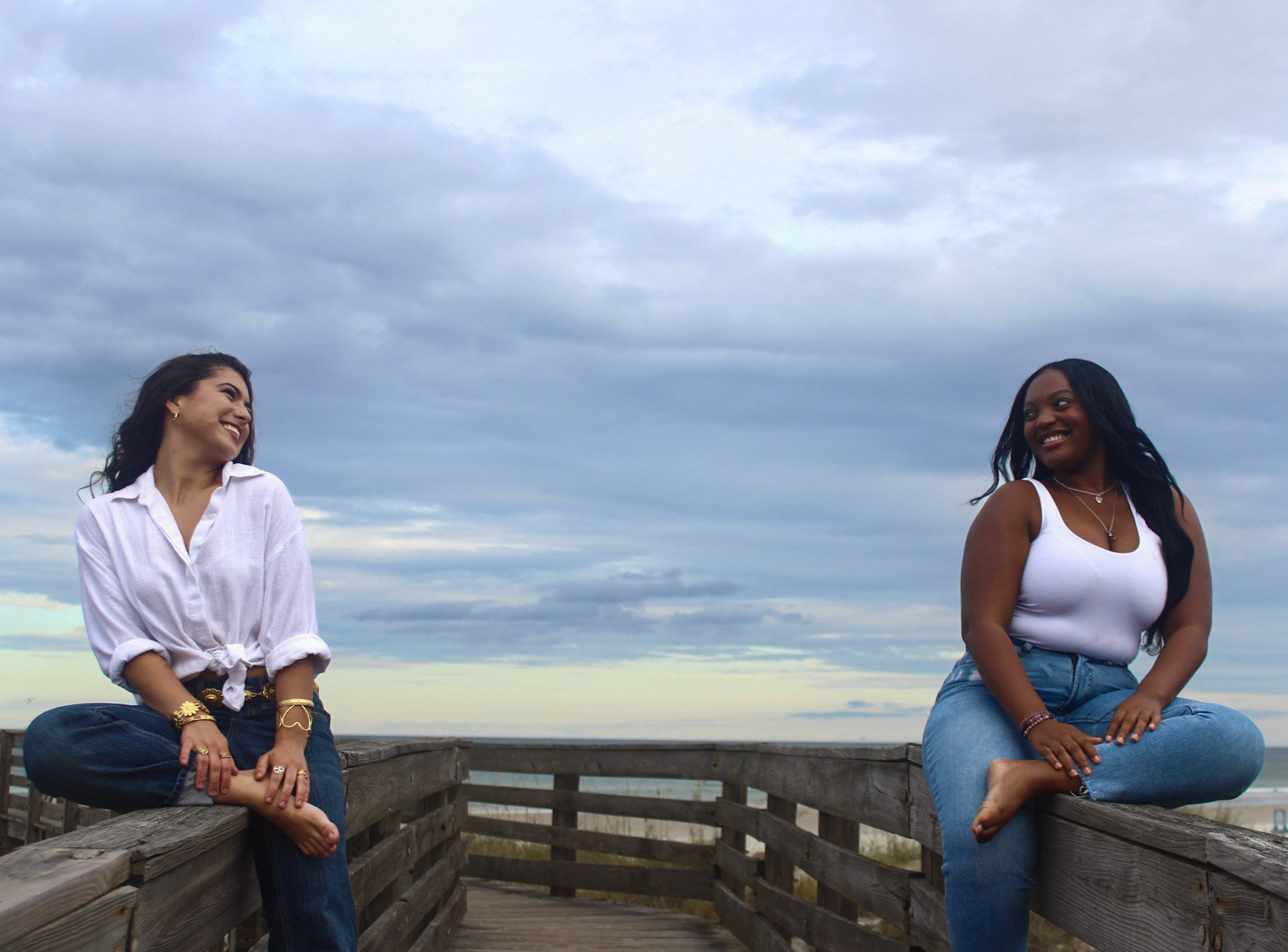 Two women sitting on a wooden railing on a beachside boardwalk, smiling and looking at each other, with overcast skies overhead.