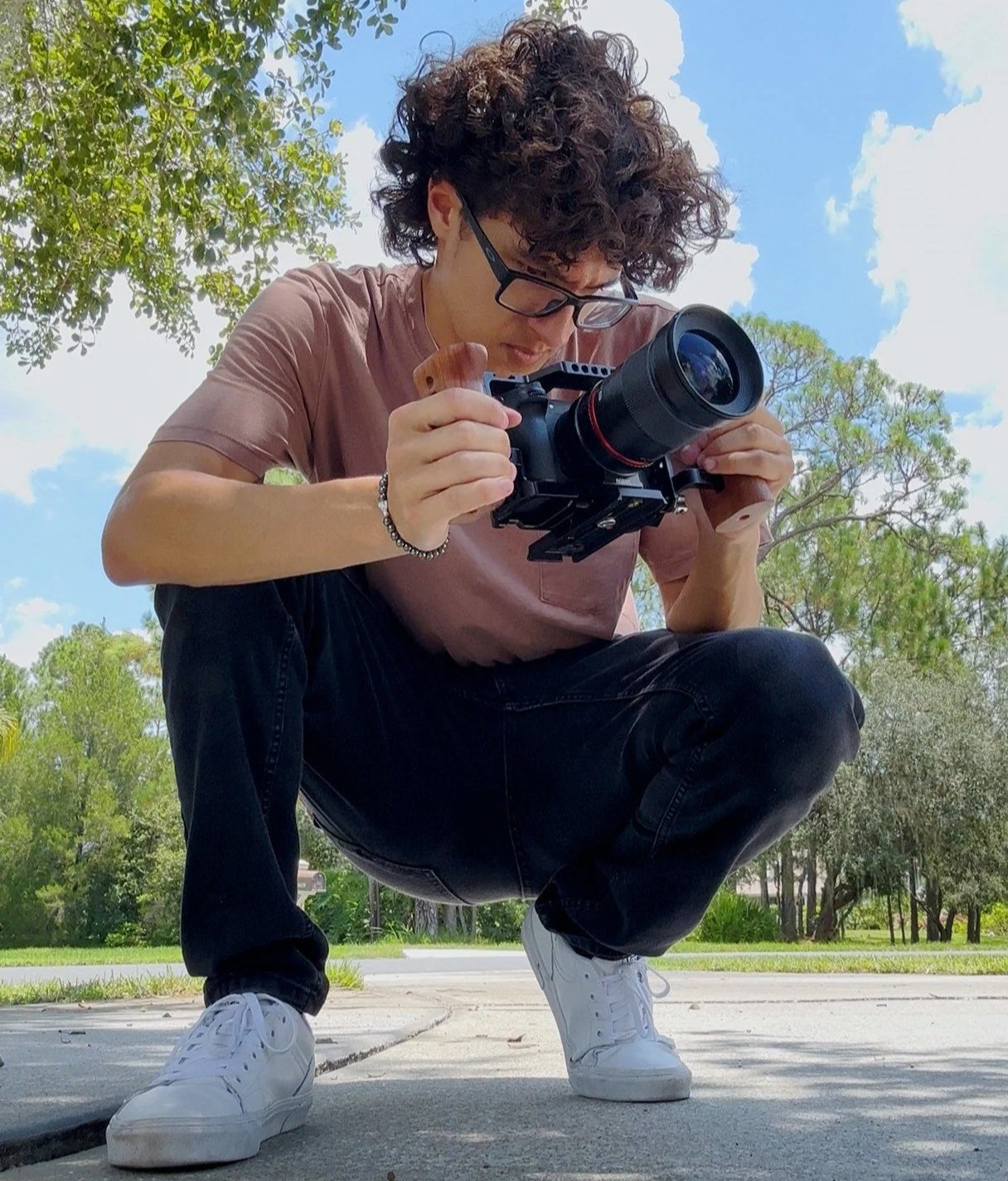 Person crouching on a sidewalk, adjusting a camera with a large lens, outdoors on a sunny day with trees and blue sky in the background.